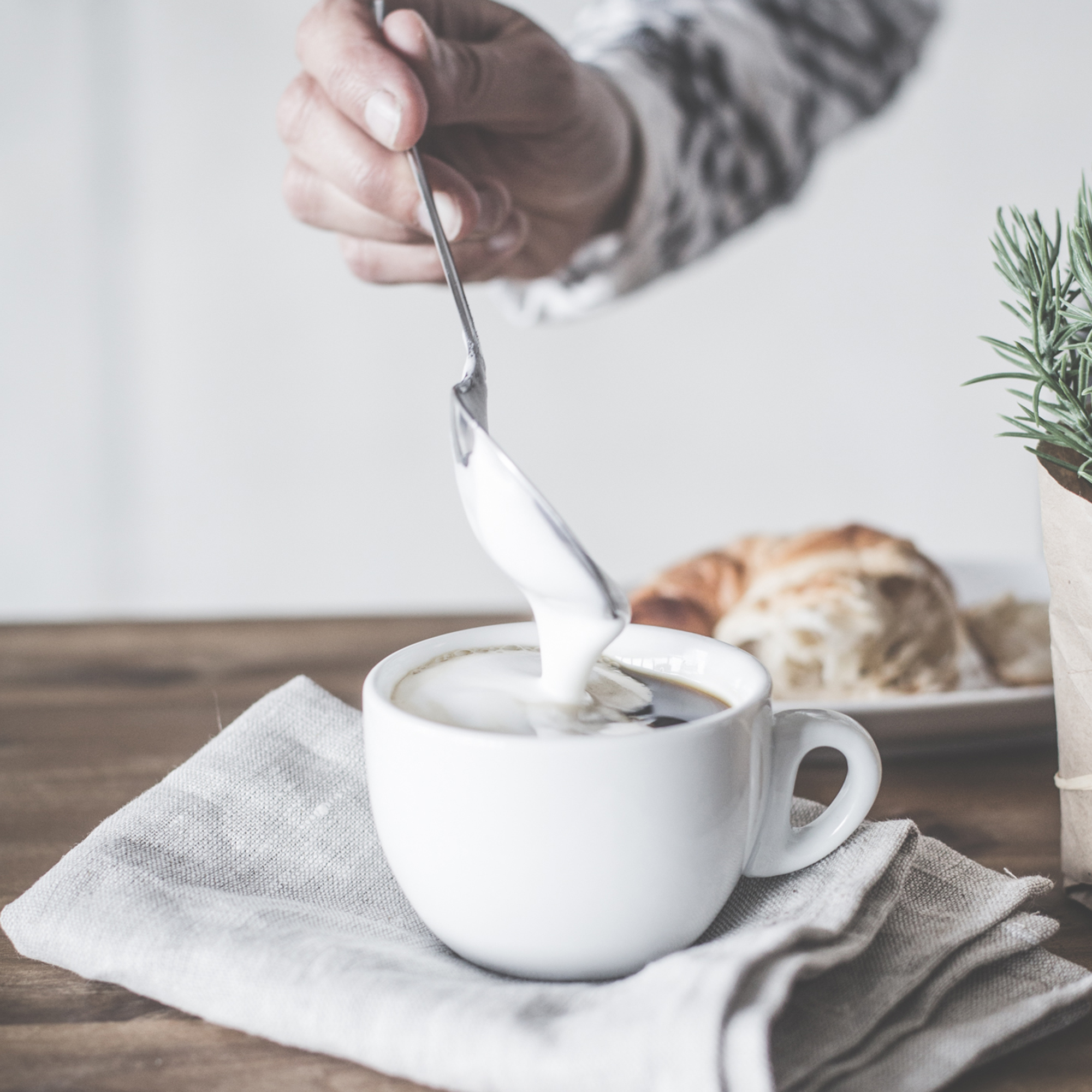 Eine Person gießt Milch in eine weiße Tasse Kaffee, mit Croissant und Pflanze.