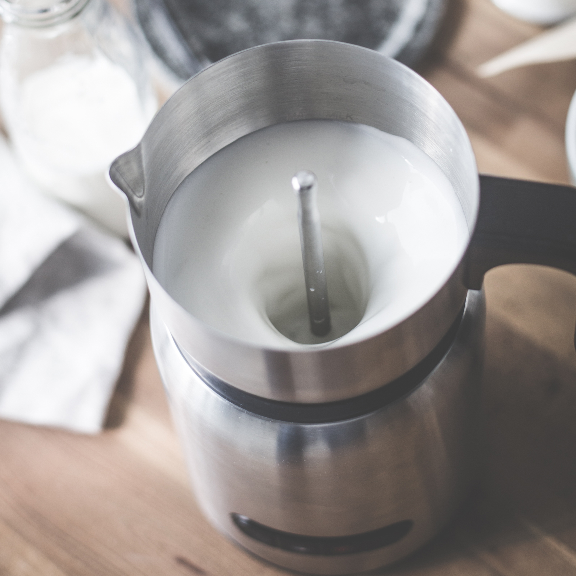 Milchaufschäumer aus Edelstahl mit Milch. Schwarzer Griff sichtbar. Holzoberfläche im Hintergrund.