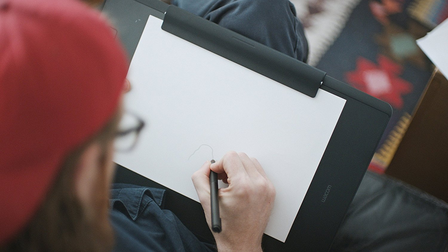 Eine Person zeichnet auf weißem Papier auf einem Tablet. Sie tragen eine rote Mütze und sitzen.