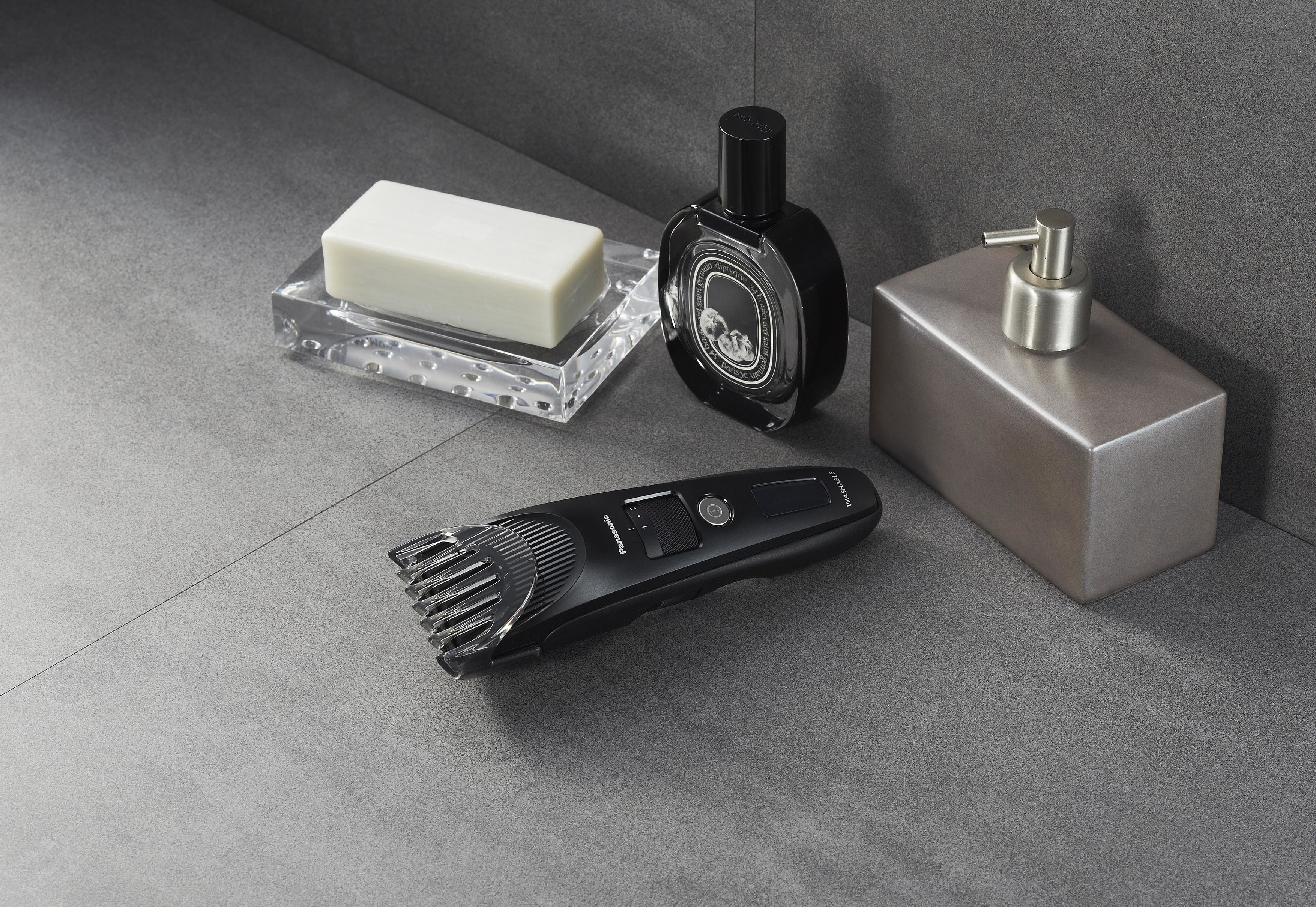Scène de salle de bain avec une tondeuse noire, du savon, du parfum et un distributeur argenté sur surface grise.