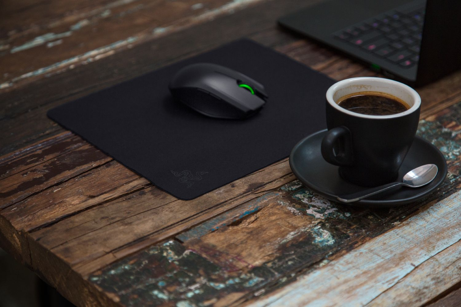 Une souris noire et une tasse de café sur une table en bois avec un ordinateur portable en arrière-plan.