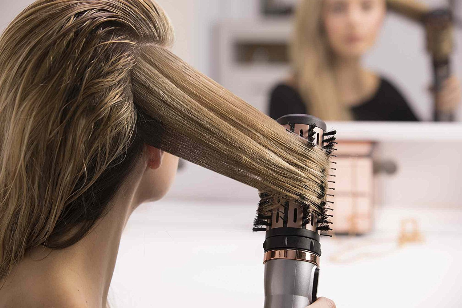 A woman styling her hair with a rotating brush. The hair is blonde and the background is blurred.