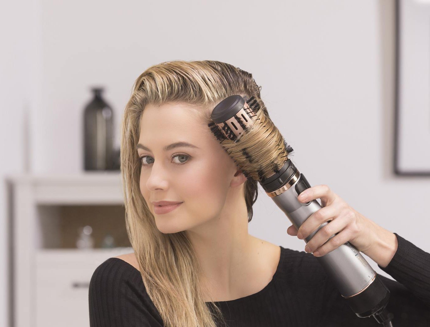 A woman using a hair dryer brush on her wet hair. She is smiling.