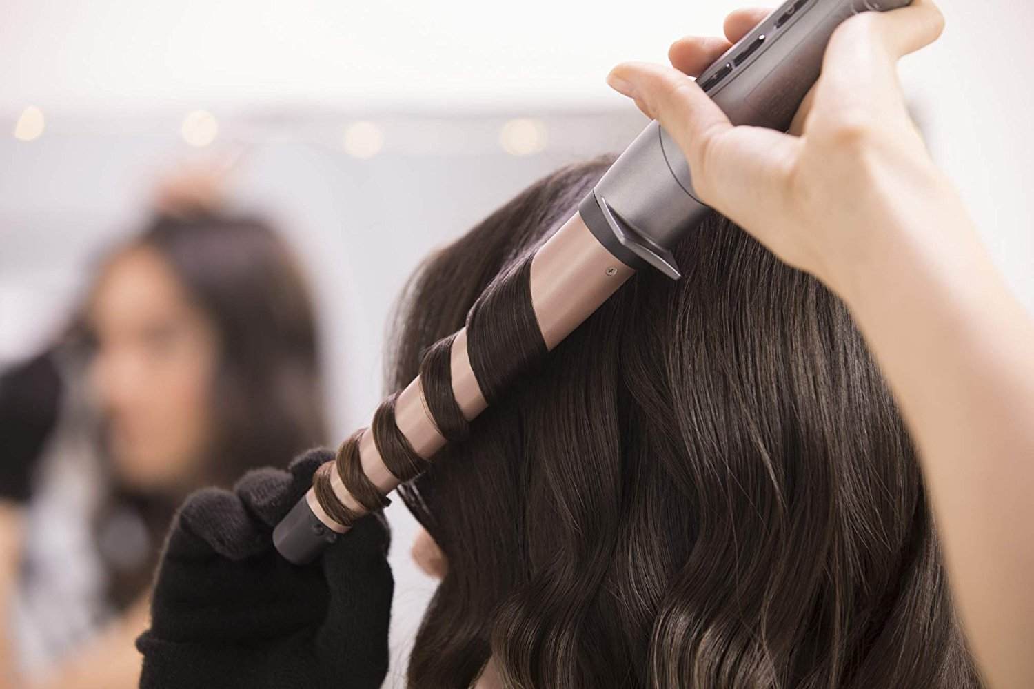 A person curling a woman's hair with a curling iron. Mirror and another person in background.