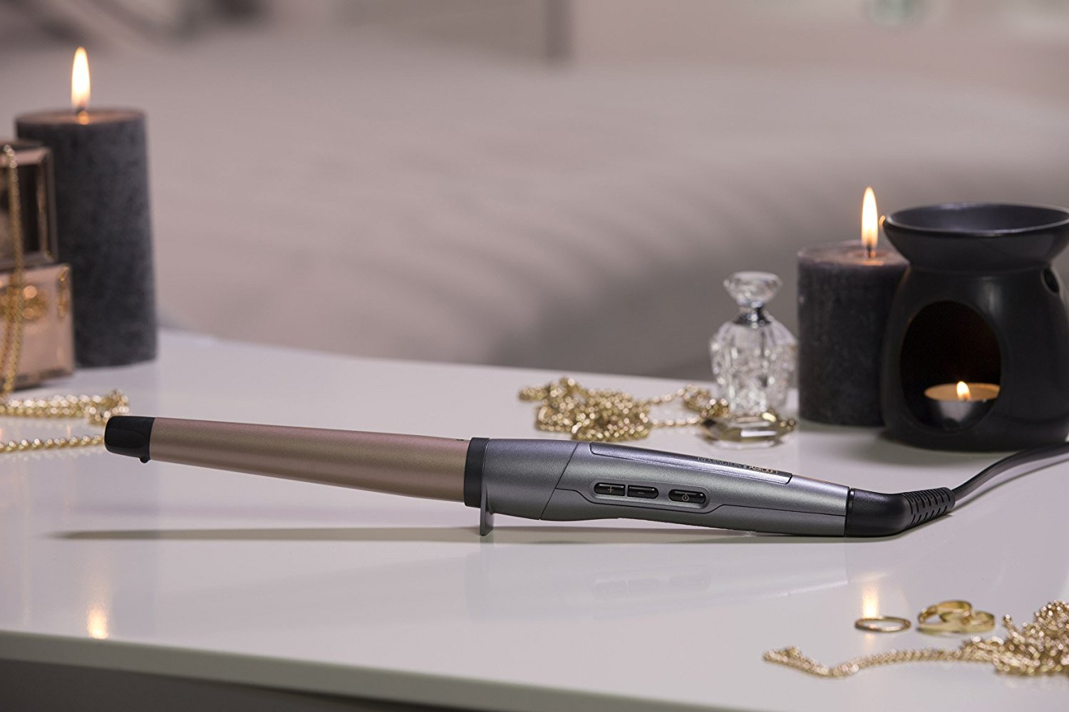 Hair curler on a table with candles, jewelry and perfume. White table, grey background.