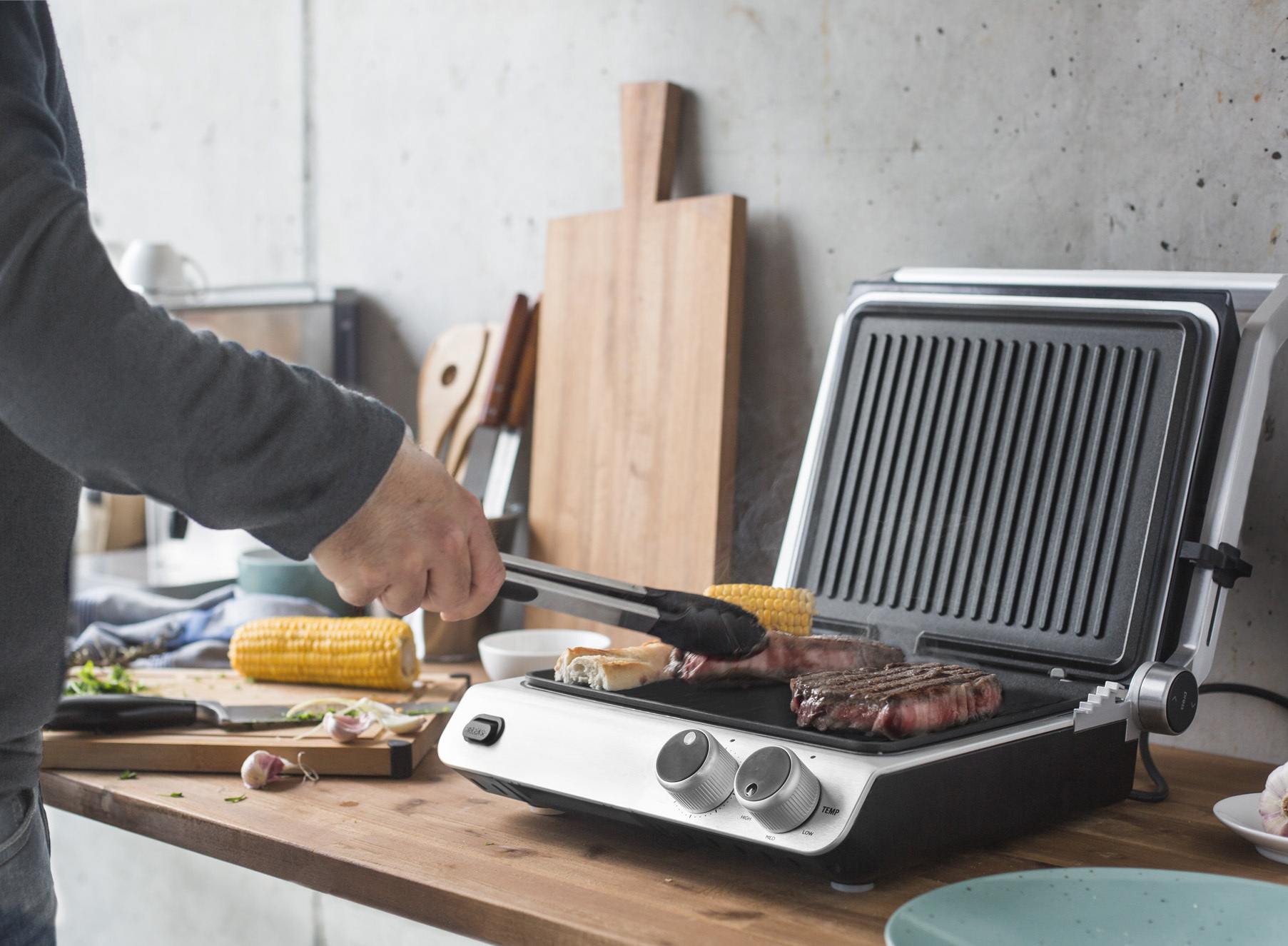 Eine Person verwendet Zangen, um Essen auf einem Grill zu wenden, Mais und andere Artikel in der Nähe.