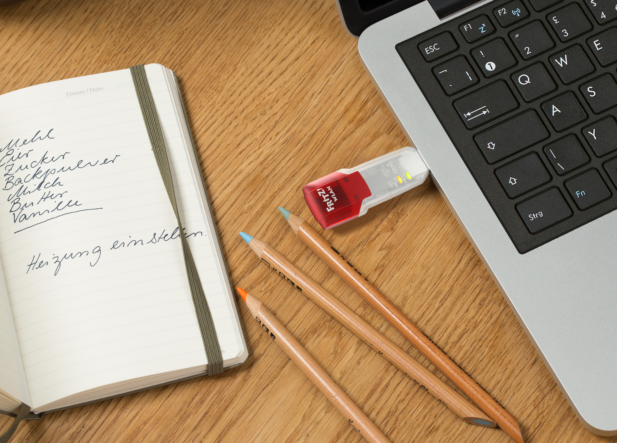 Ein Laptop mit USB-Stick und Buntstiften neben einem Notizbuch mit handschriftlichem Text.