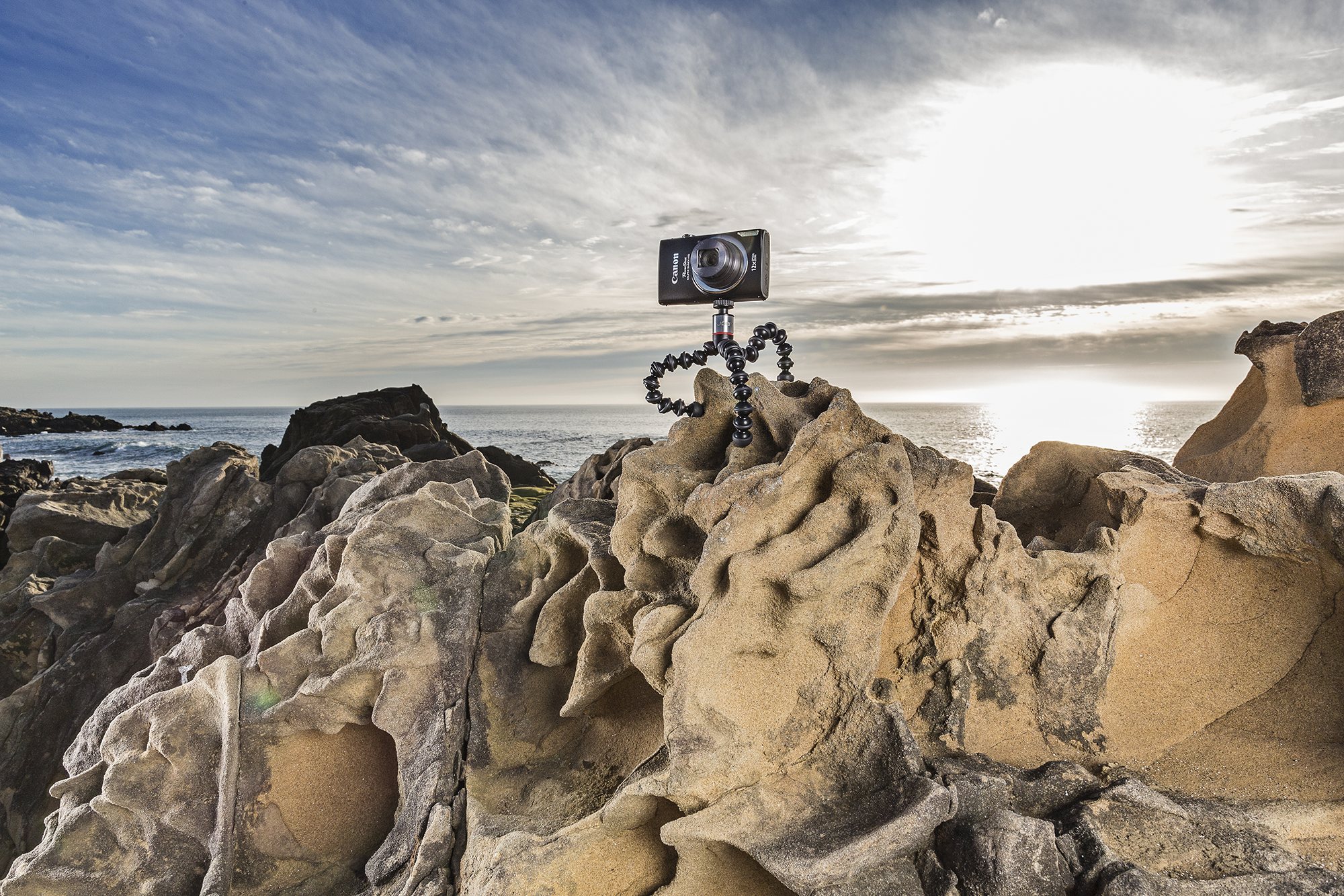 A camera on a flexible tripod on a rock formation near the ocean, with a cloudy sky.