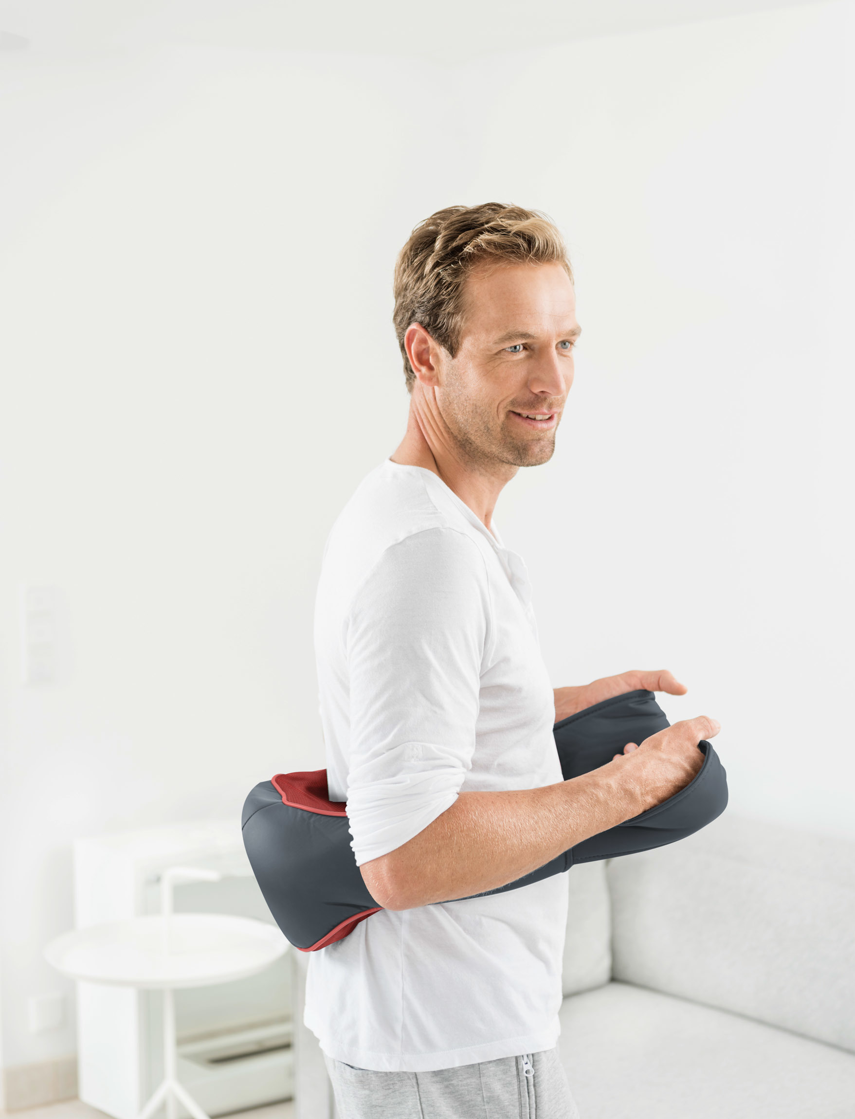 Mann lächelt, während er ein Massagegerät auf dem Rücken hält, in einer hellen Raumumgebung.