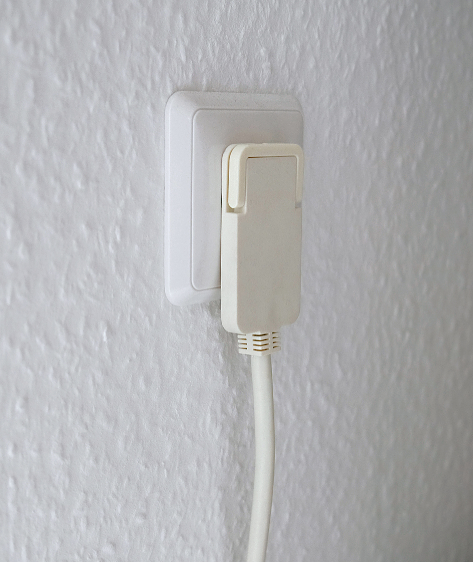 Weiße Steckdose mit eingestecktem weißen Stecker. Die Wand ist strukturiert und weiß gestrichen.