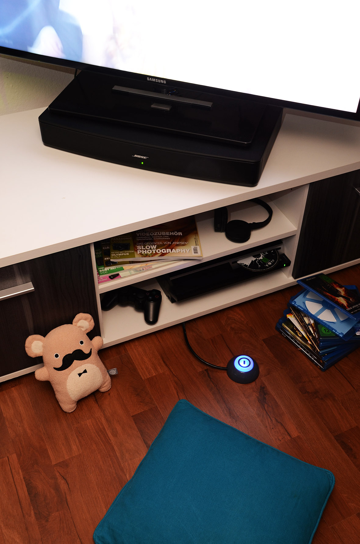 Wohnzimmer mit Fernseher, Soundanlage, Spielzeug und Fernbedienung. Holzboden und blaues Kissen.
