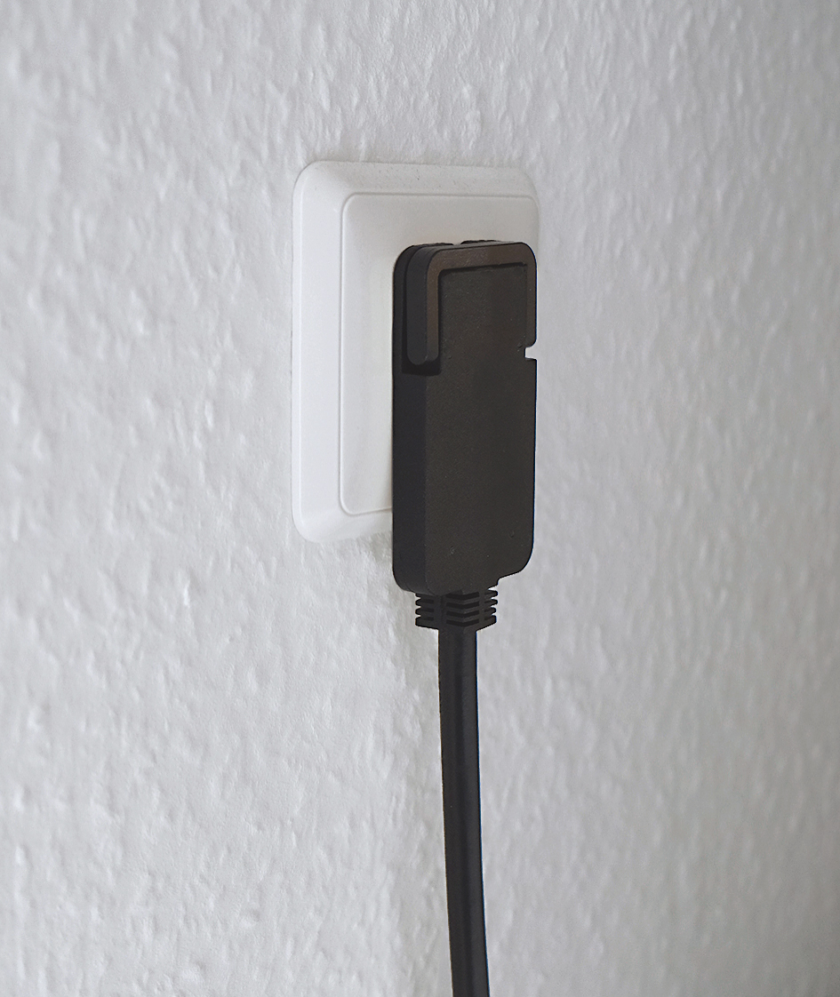 Ein schwarzer Netzadapter ist in eine weiße Steckdose an einer weißen, strukturierten Wand gesteckt.