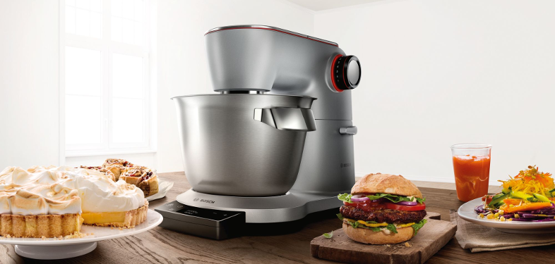 Egy Bosch konyhai robotgép ételekkel az asztalon: sütemény, pékáruk, hamburger, saláta és gyümölcslé.