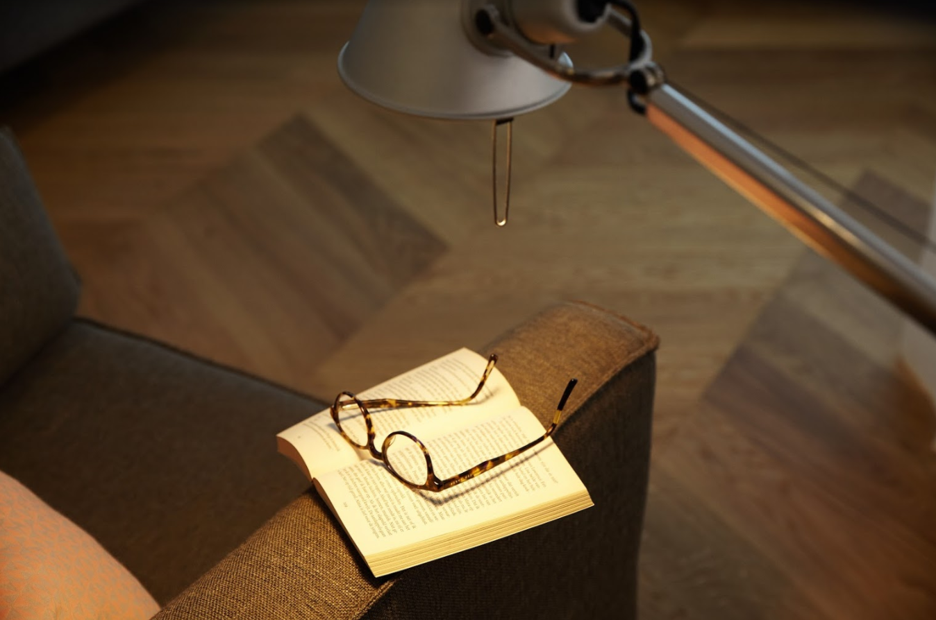 Open boek met bril op een fauteuil, onder een lamp. Houten vloer op de achtergrond.
