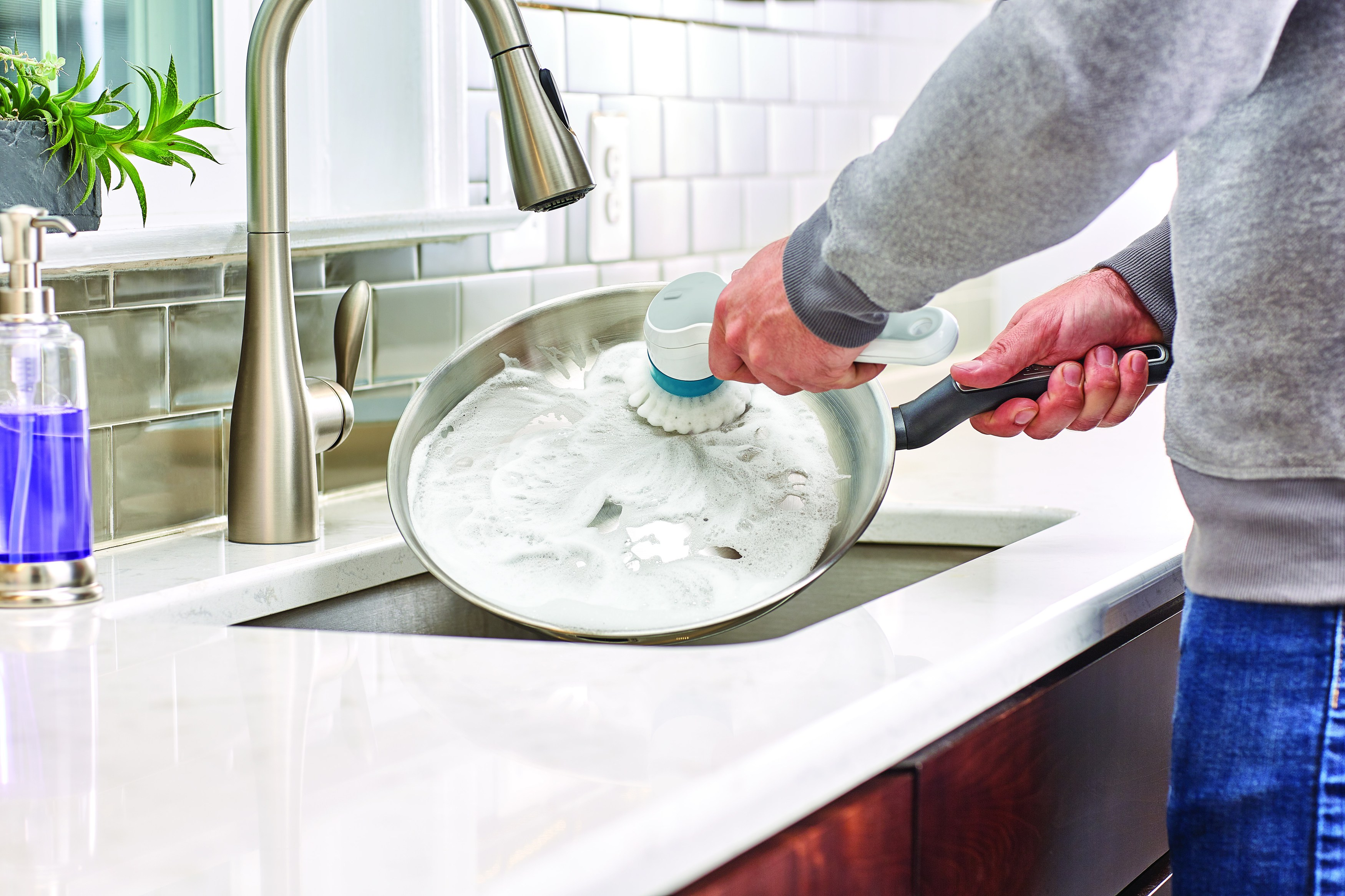 Eine Person wäscht eine Pfanne in einem Waschbecken mit einer Bürste und Seifenblasen. Küchenumgebung, Fokus auf die Reinigung.