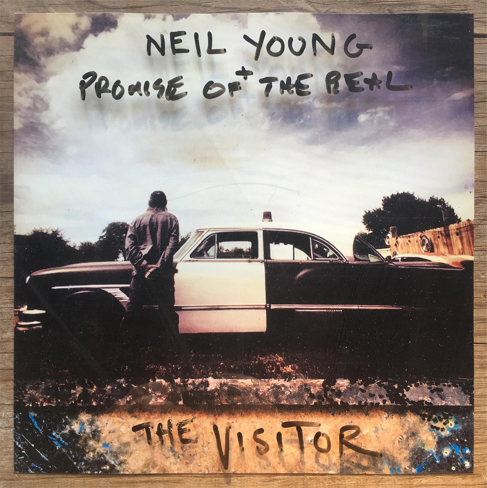 Albumhoes van Neil Youngs The Visitor. Een man staat bij een auto. Lucht en bomen op de achtergrond.