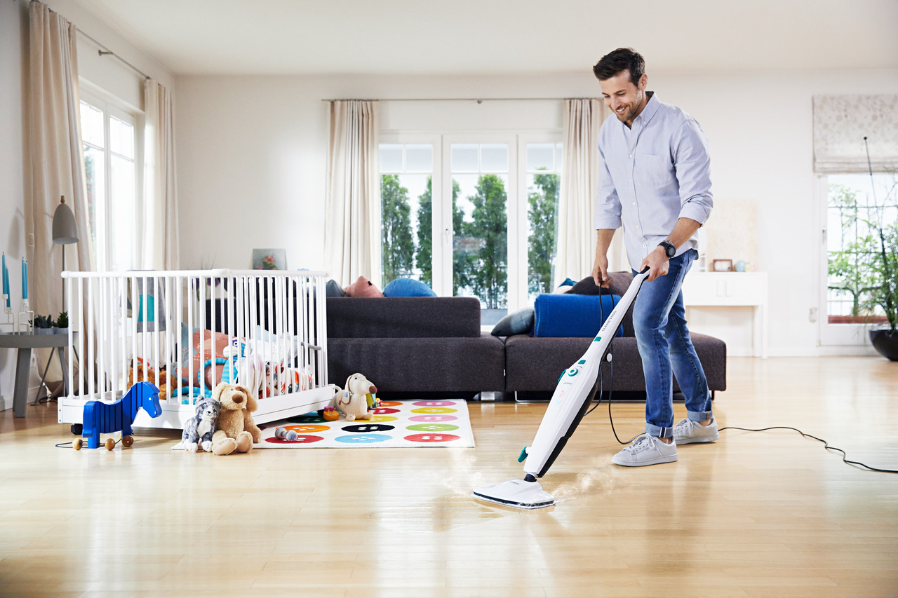 Mann saugt Wohnzimmer. Babybett und Spielzeug sind im Hintergrund.