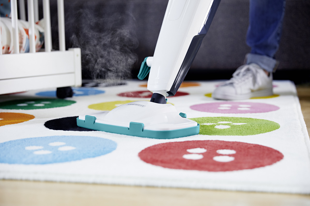 Ein Dampfreiniger reinigt einen bunten Teppich mit Knopfpflasterung. Im Hintergrund ist ein Kinderbett.