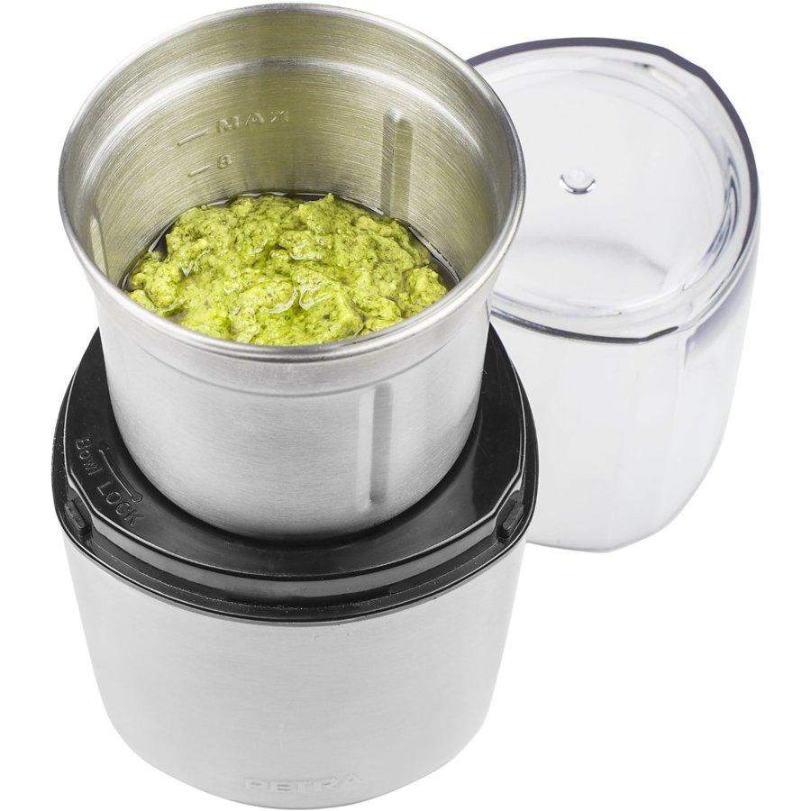 A stainless steel food processor with green pesto inside, and a clear lid beside it on a white surface.