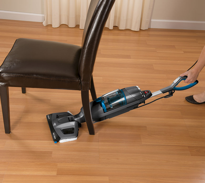 Un aspirateur nettoie un sol. Il y a une chaise en arrière-plan. L'aspirateur est noir et gris.