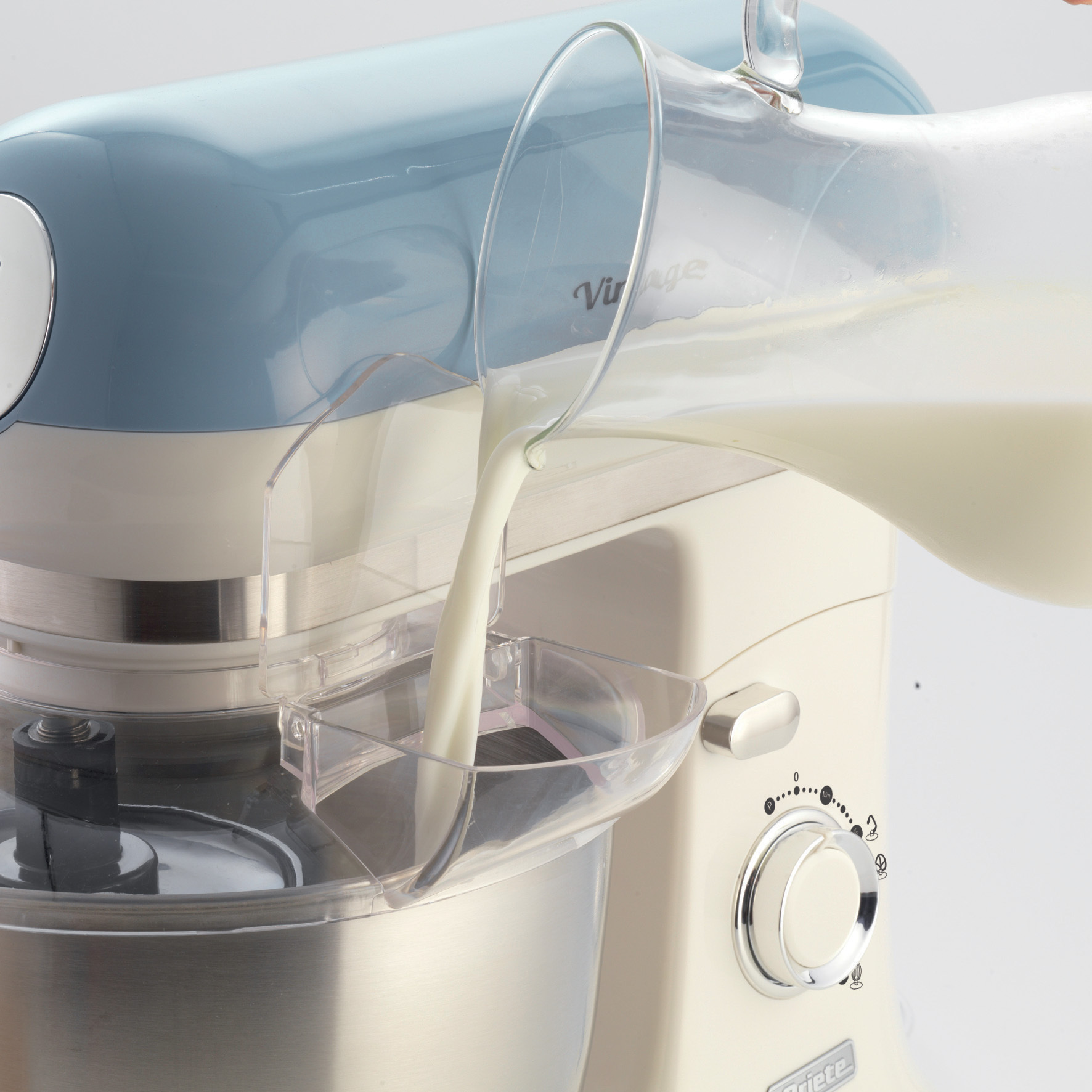 Un mixer da cucina con latte versato nella ciotola da una brocca trasparente, di colore blu e crema.