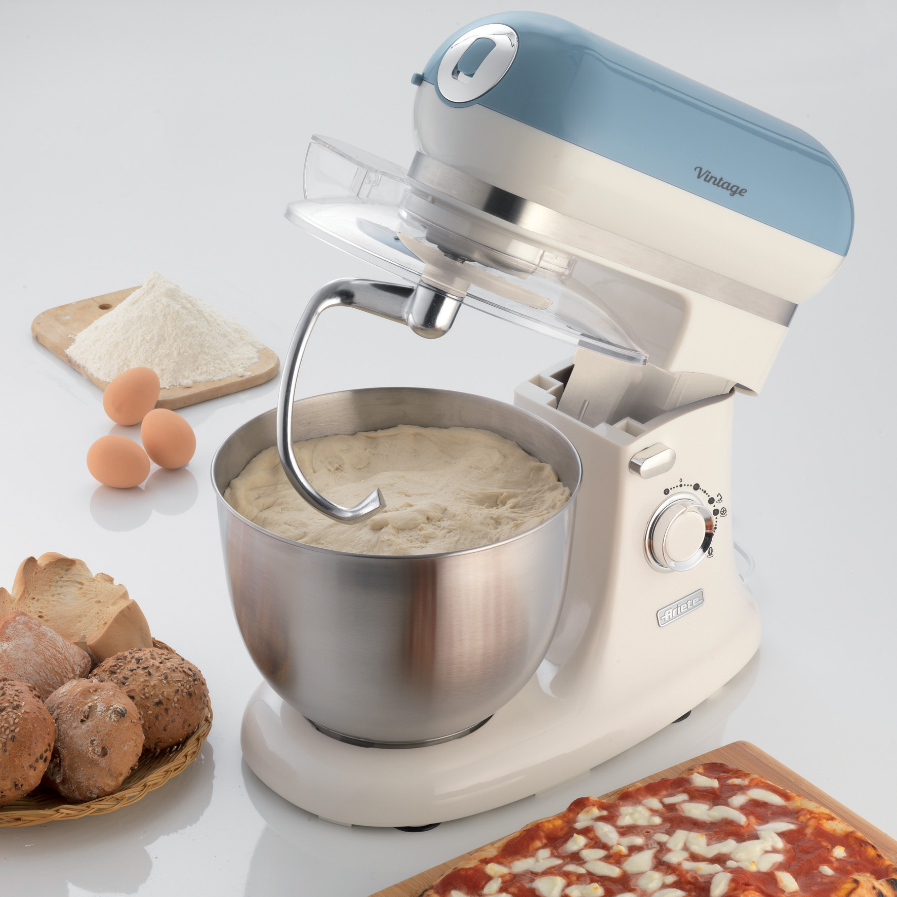 Un'impastatrice in stile vintage con ingredienti intorno, tra cui farina, uova e pane. Sfondo bianco.