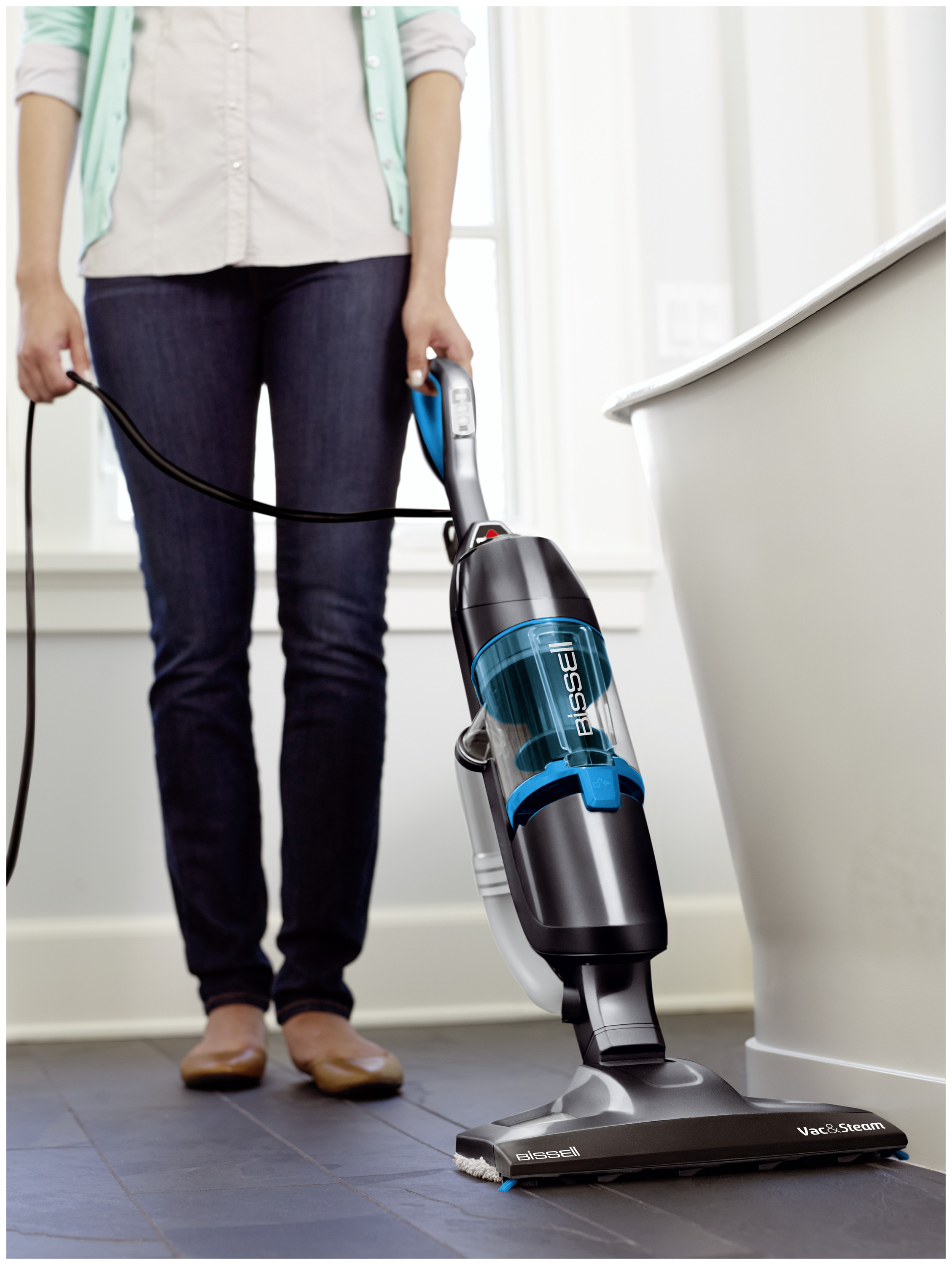 Eine Frau benutzt einen Bissell Vac & Steam Reiniger auf einem dunklen Boden, im Hintergrund eine weiße Wand.