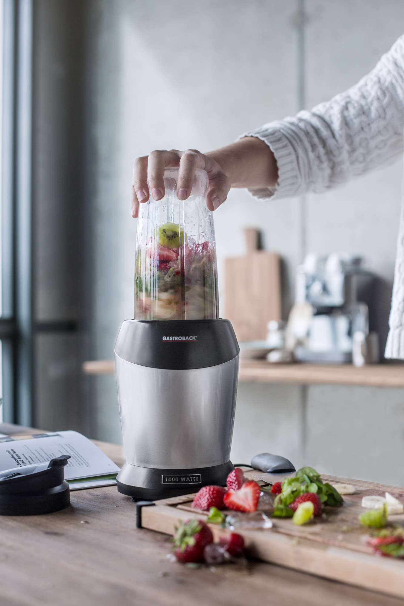 Eine Person, die einen Mixer mit Obst auf einem Holztisch benutzt.