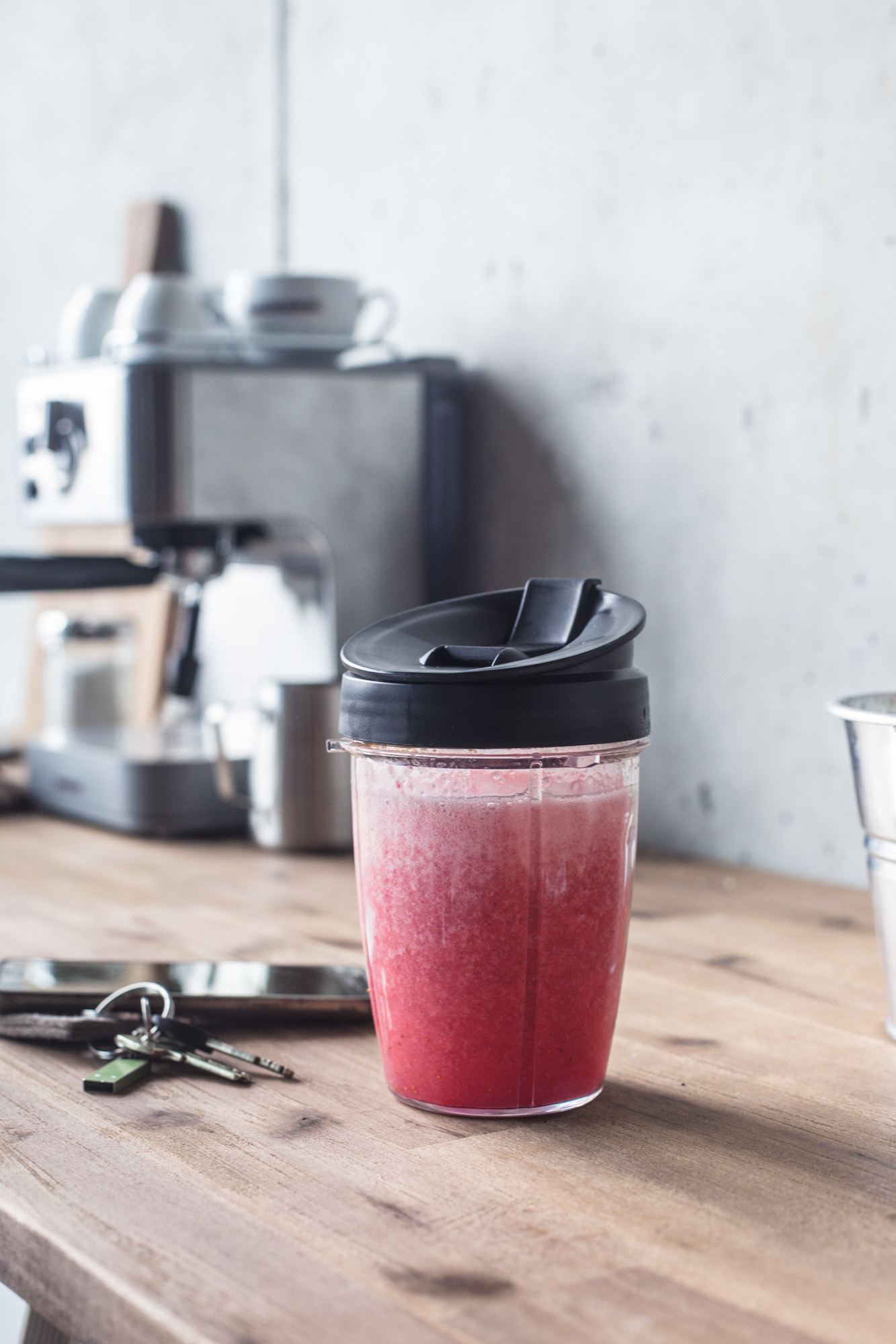 Ein Smoothie in einem durchsichtigen Becher mit schwarzem Deckel, mit Schlüsseln und Kaffeemaschine auf einem Holztisch.