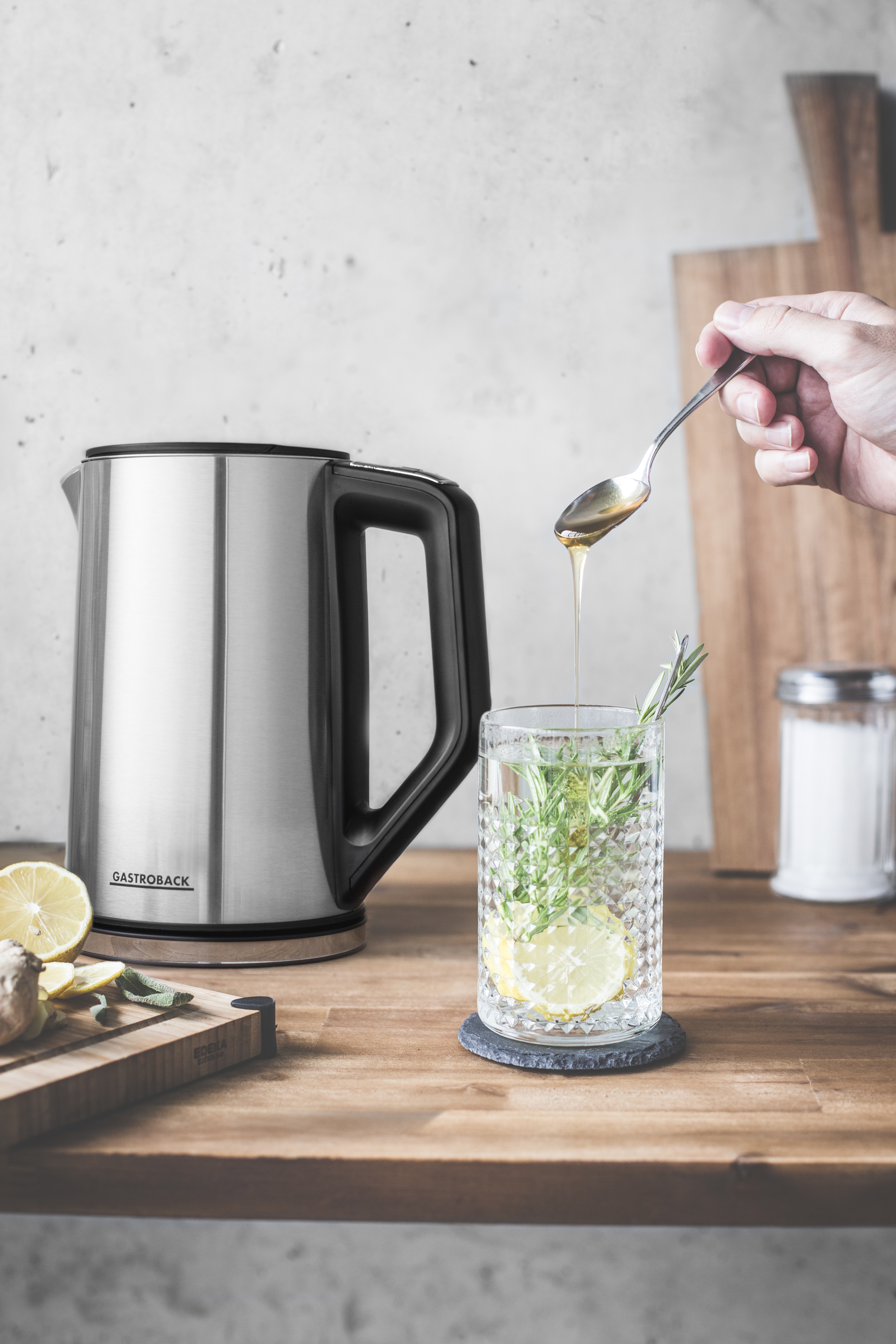 Person gießt Honig in Glas mit Zitrone und Rosmarin, neben Wasserkocher.