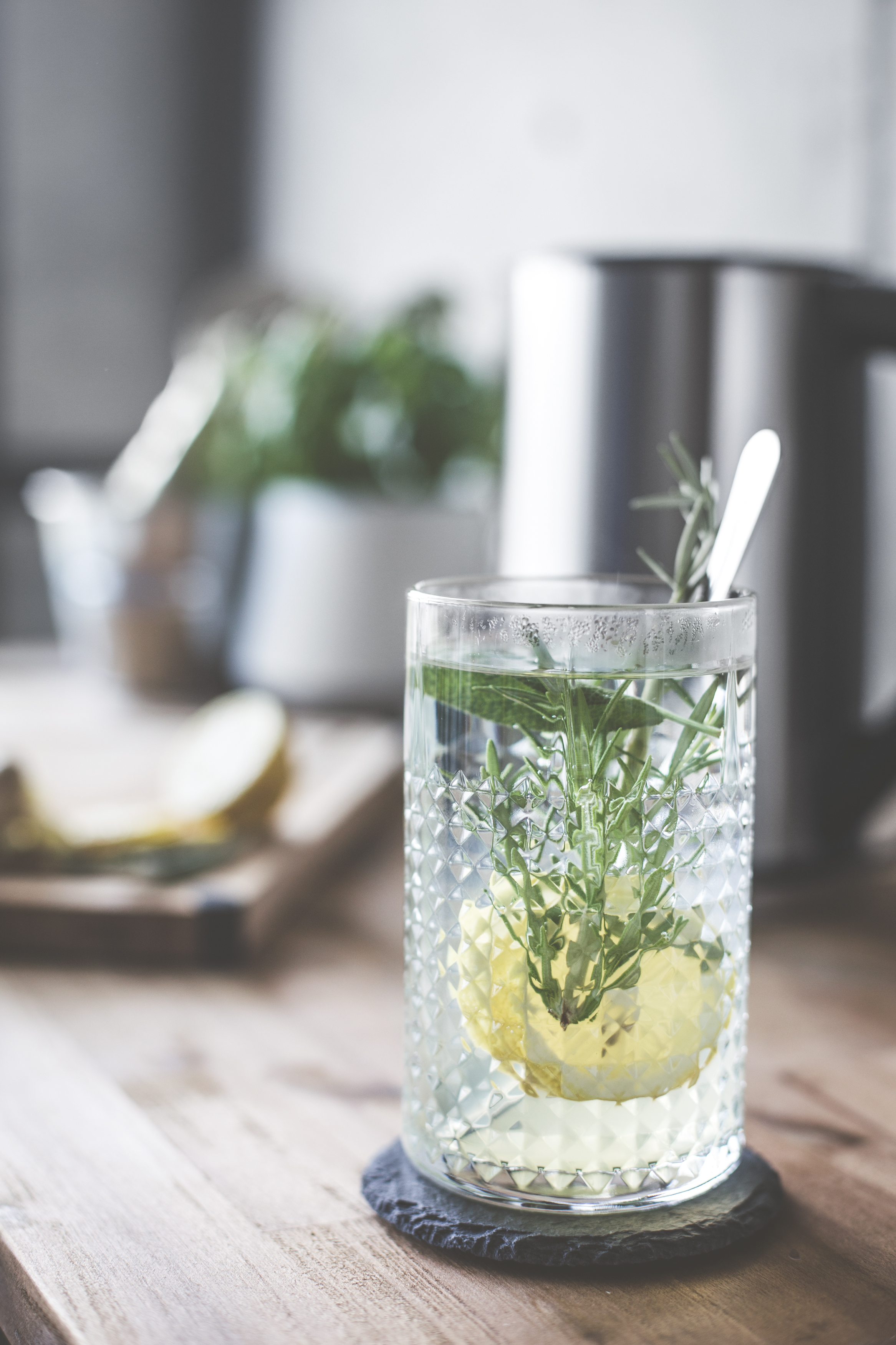 Glas mit Wasser, Zitrone und Rosmarin. Teekessel im Hintergrund.