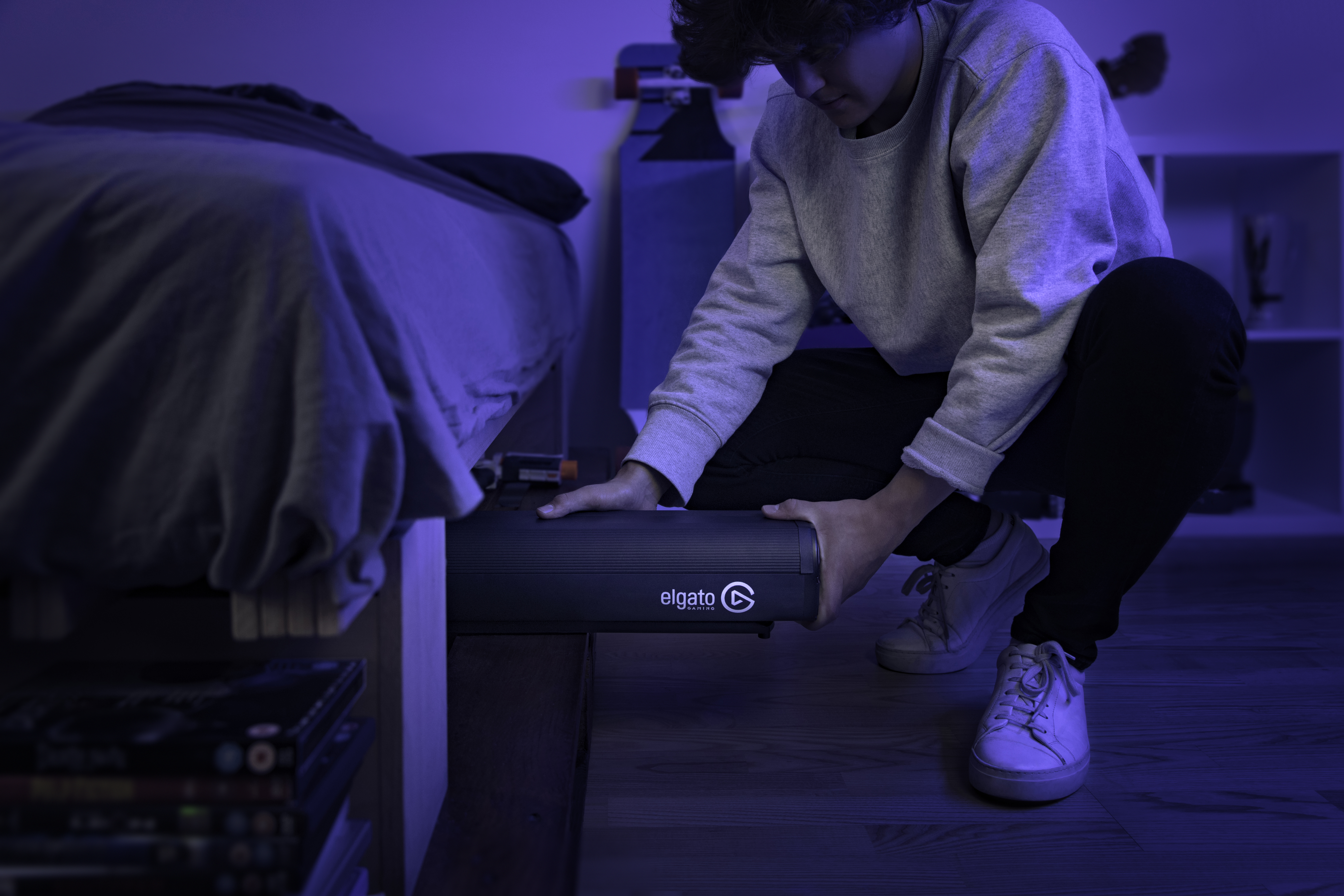 Eine Person, die einen schwarzen Lautsprecher unter ein Bett in einem blau beleuchteten Raum legt.