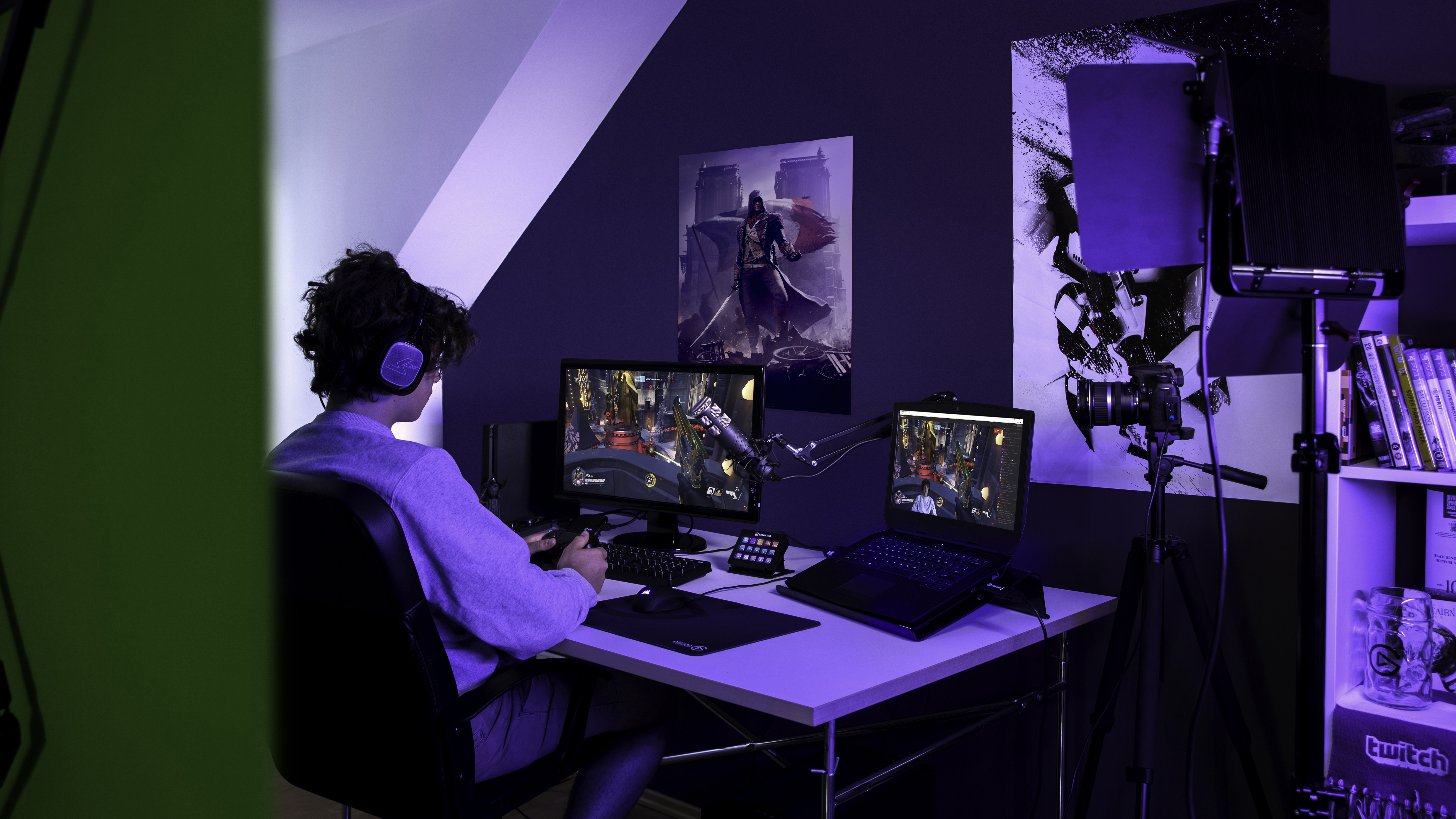 Eine Person spielt an einem Schreibtisch mit Monitor, Laptop und Spielkonsole in einem Raum.
