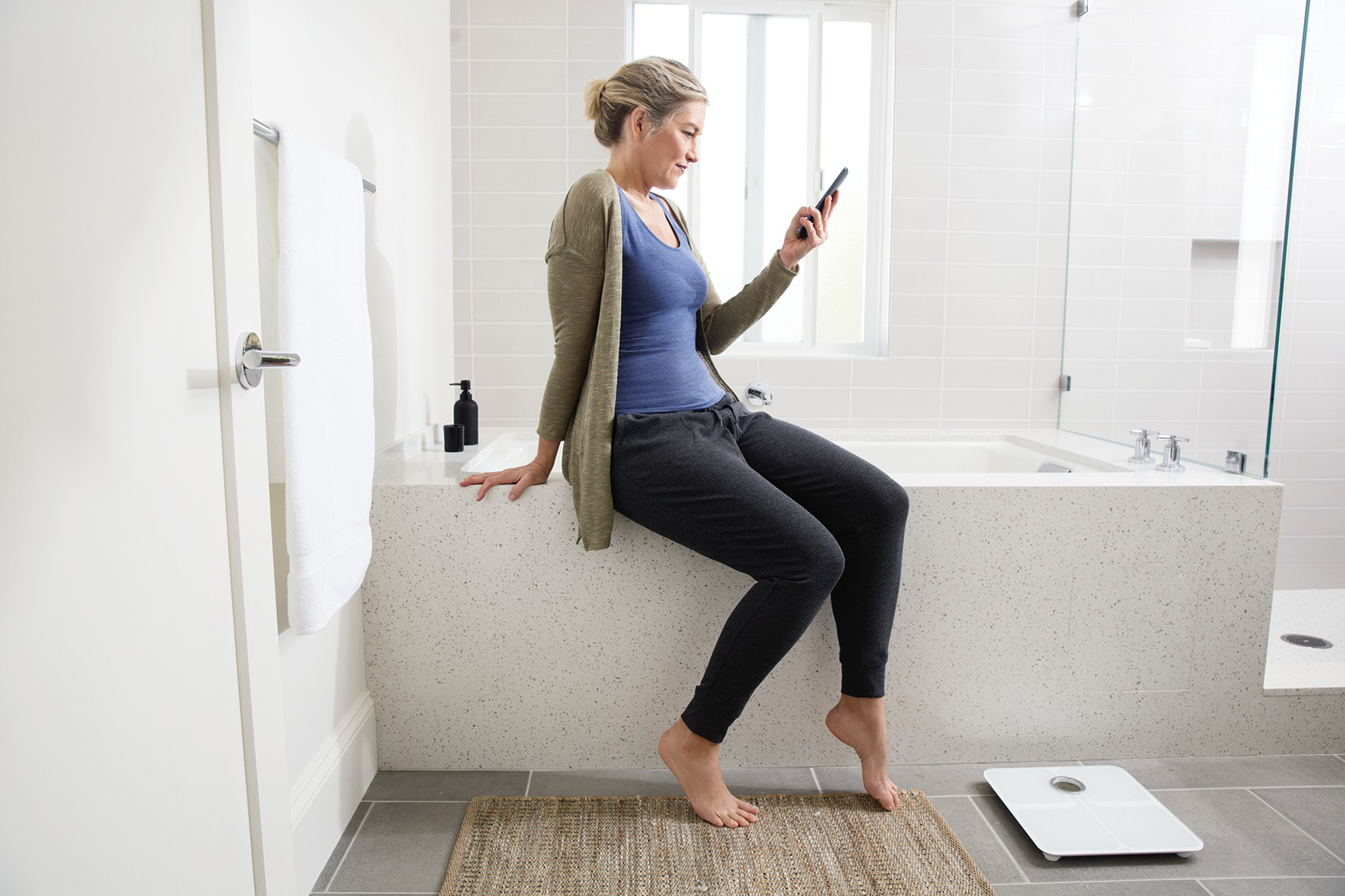 Frau im Badezimmer, die auf einer Theke sitzt und auf ihr Telefon schaut, Waage auf dem Boden.