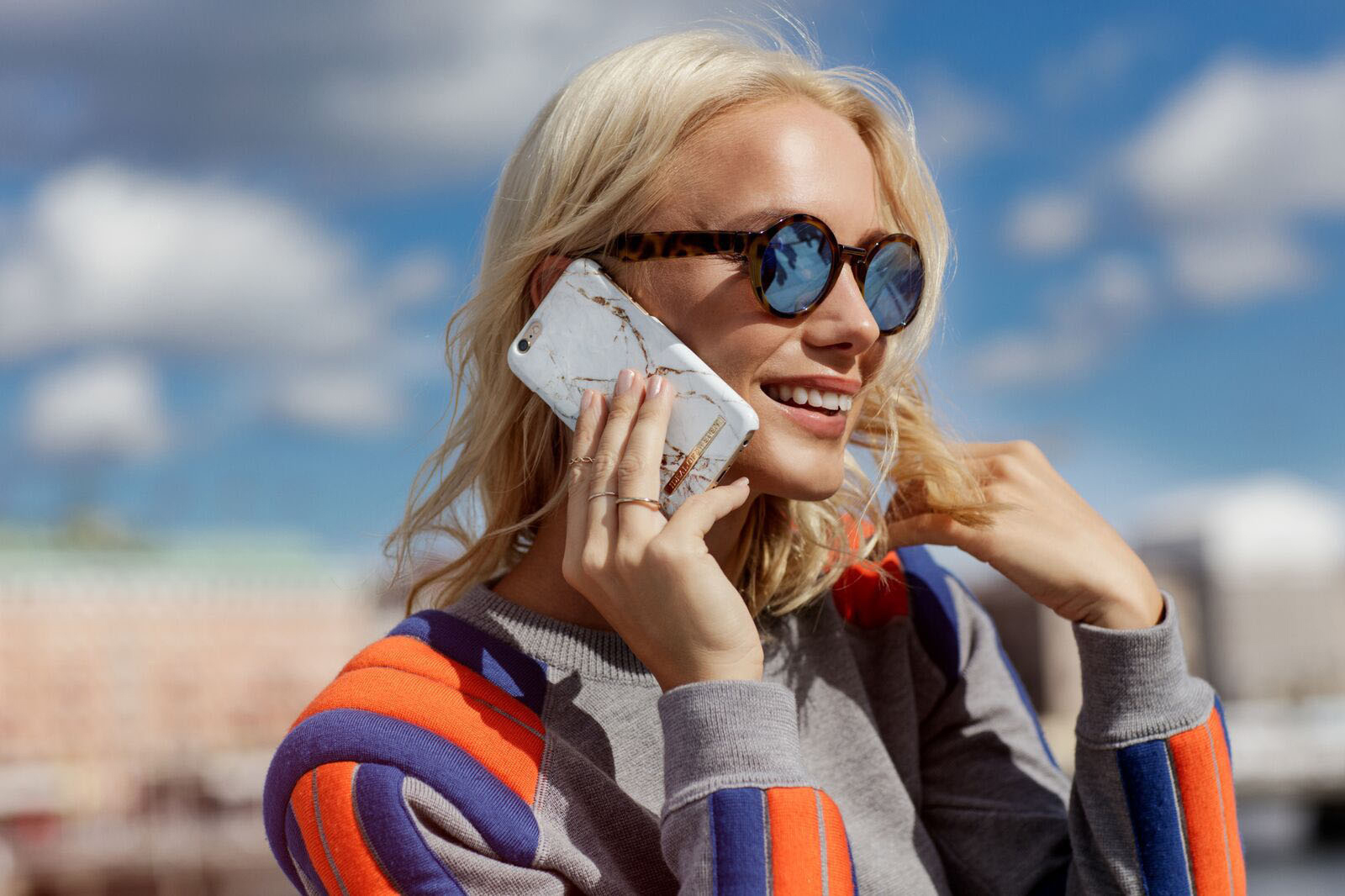 Frau mit Sonnenbrille, telefoniert. Trägt farbenfrohes Oberteil, blauer Himmel im Hintergrund.