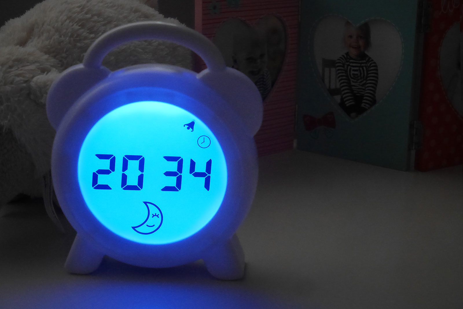 Witte wekker met een blauw verlicht digitaal display, met tijd 20:34. Andere items op de achtergrond.