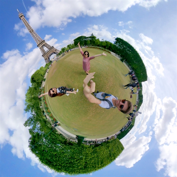 A fisheye view shows the Eiffel Tower and three people. One lies on grass, two stand with arms outstretched. Blue sky with clouds.