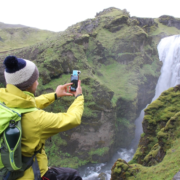 Man taking a photo of a waterfall with a phone. He wears a yellow jacket and a hat, with a green backpack. Lush green mountains.