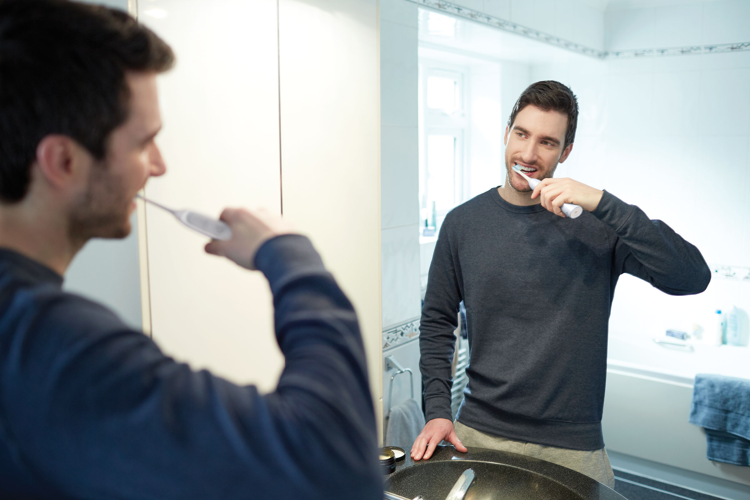 Mann putzt sich im Badezimmer die Zähne, im Spiegel reflektiert. Weiße Zahnbürste in der Hand.