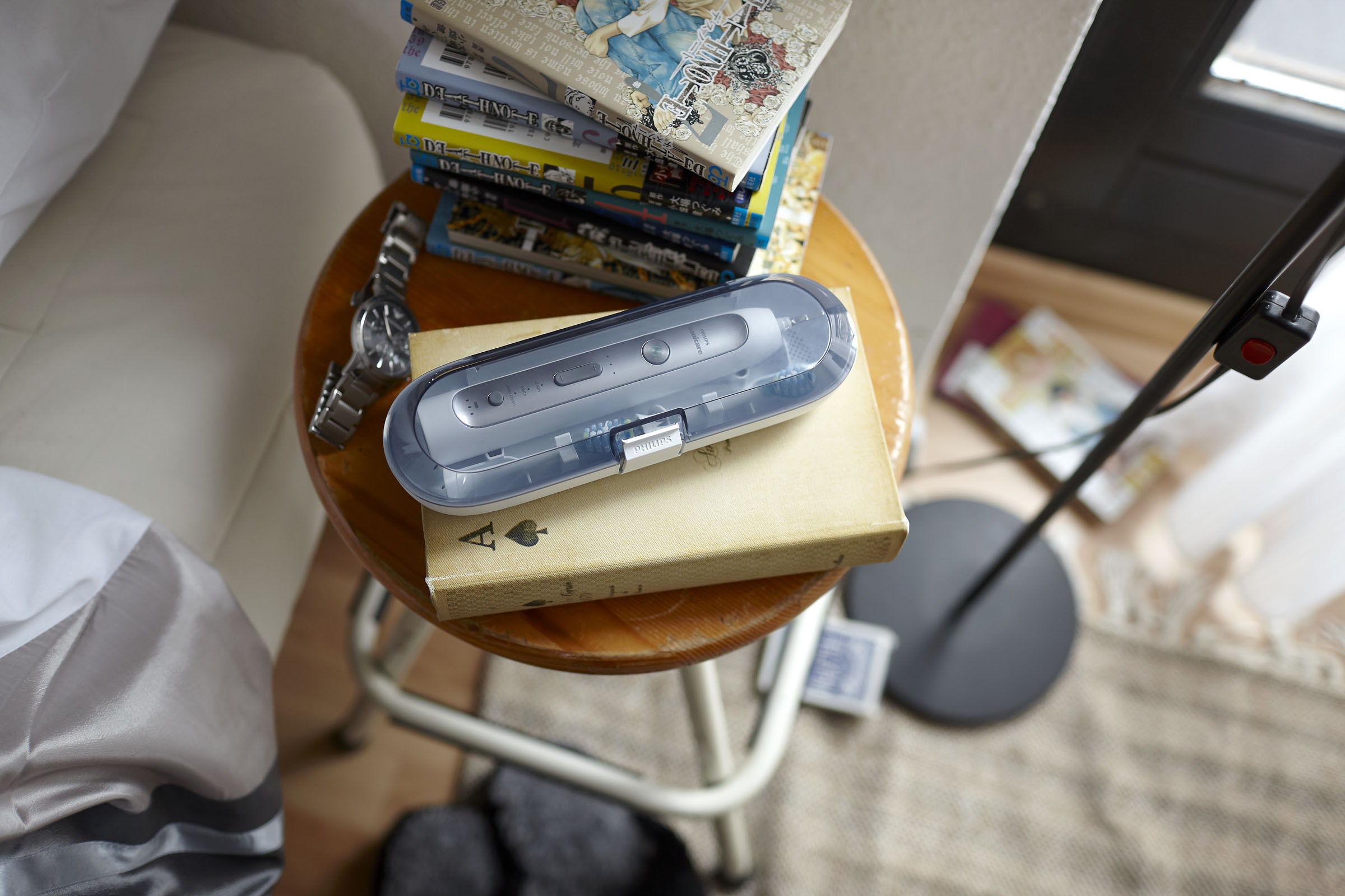 Zahnbürstenetui auf einem kleinen Tisch mit Büchern und einer Uhr. Hintergrund: ein Bett und ein Teppich.
