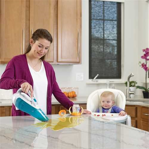 A woman vacuuming a spilled liquid on a countertop near a baby in a high chair.