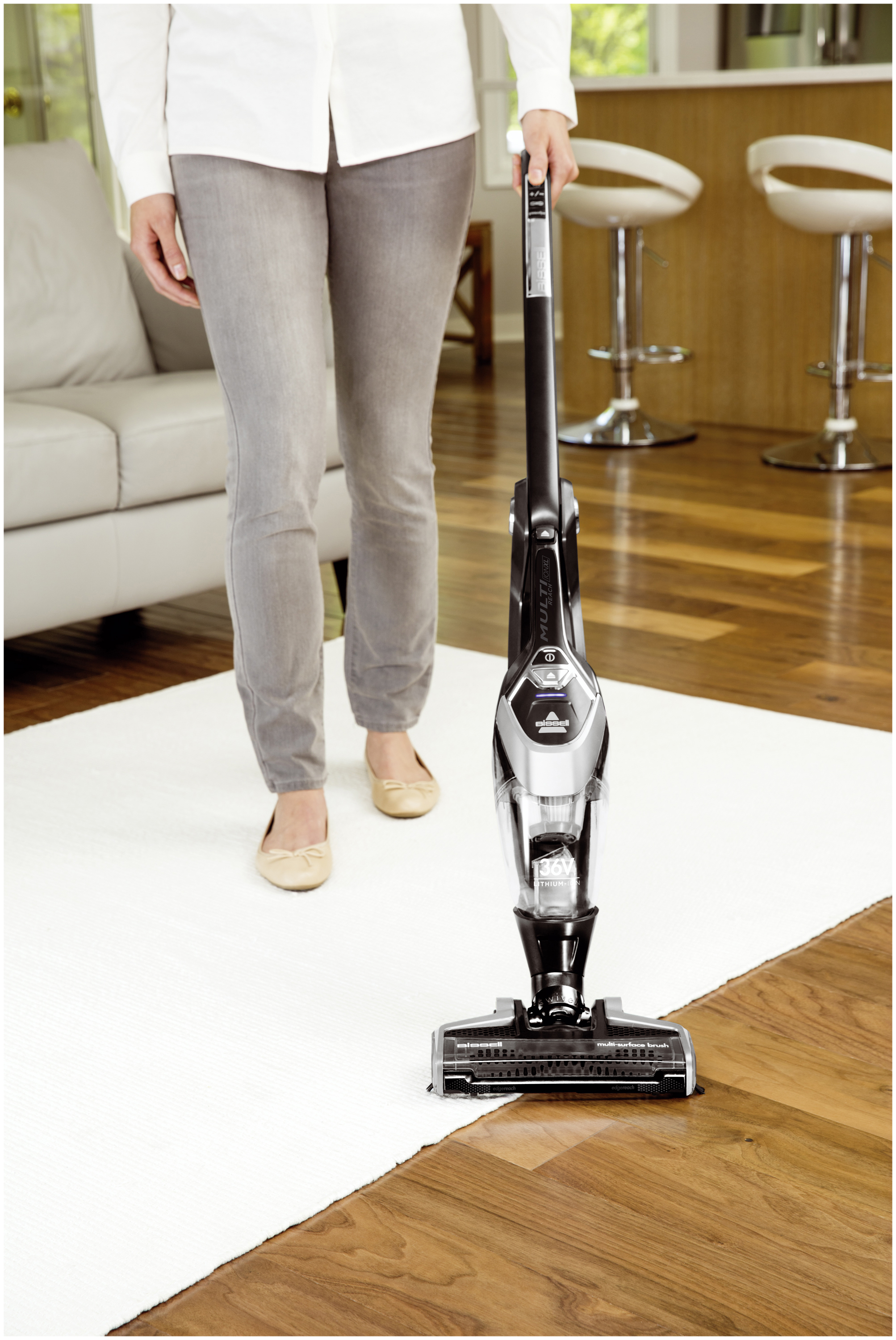 Eine Person saugt einen weißen Teppich mit einem schwarzen Staubsauger. Der Hintergrund zeigt ein Sofa und Barhocker.