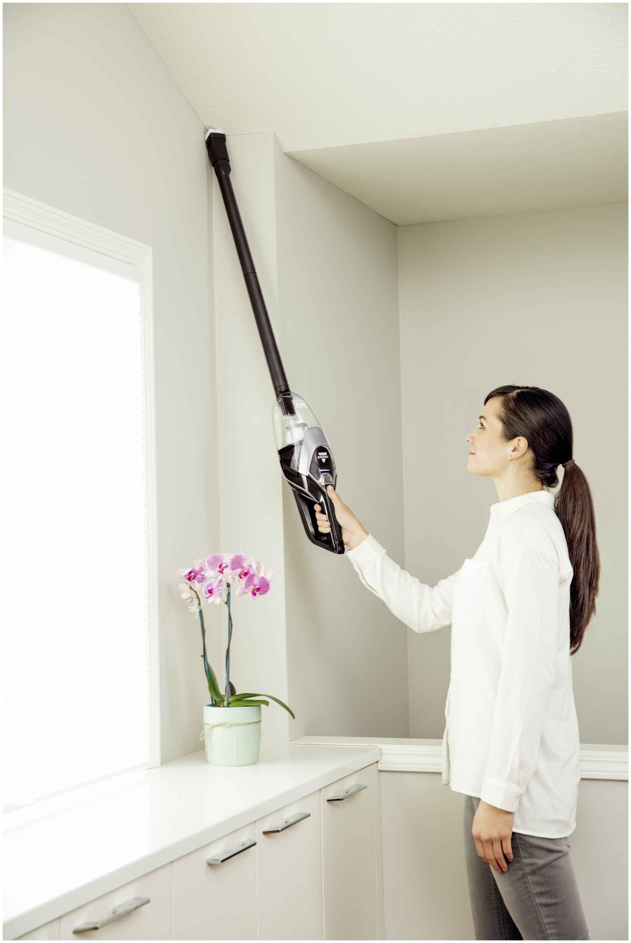Eine Frau saugt eine Wandecke mit einem Handstaubsauger. Im Hintergrund sind ein Fenster und eine Orchidee zu sehen.