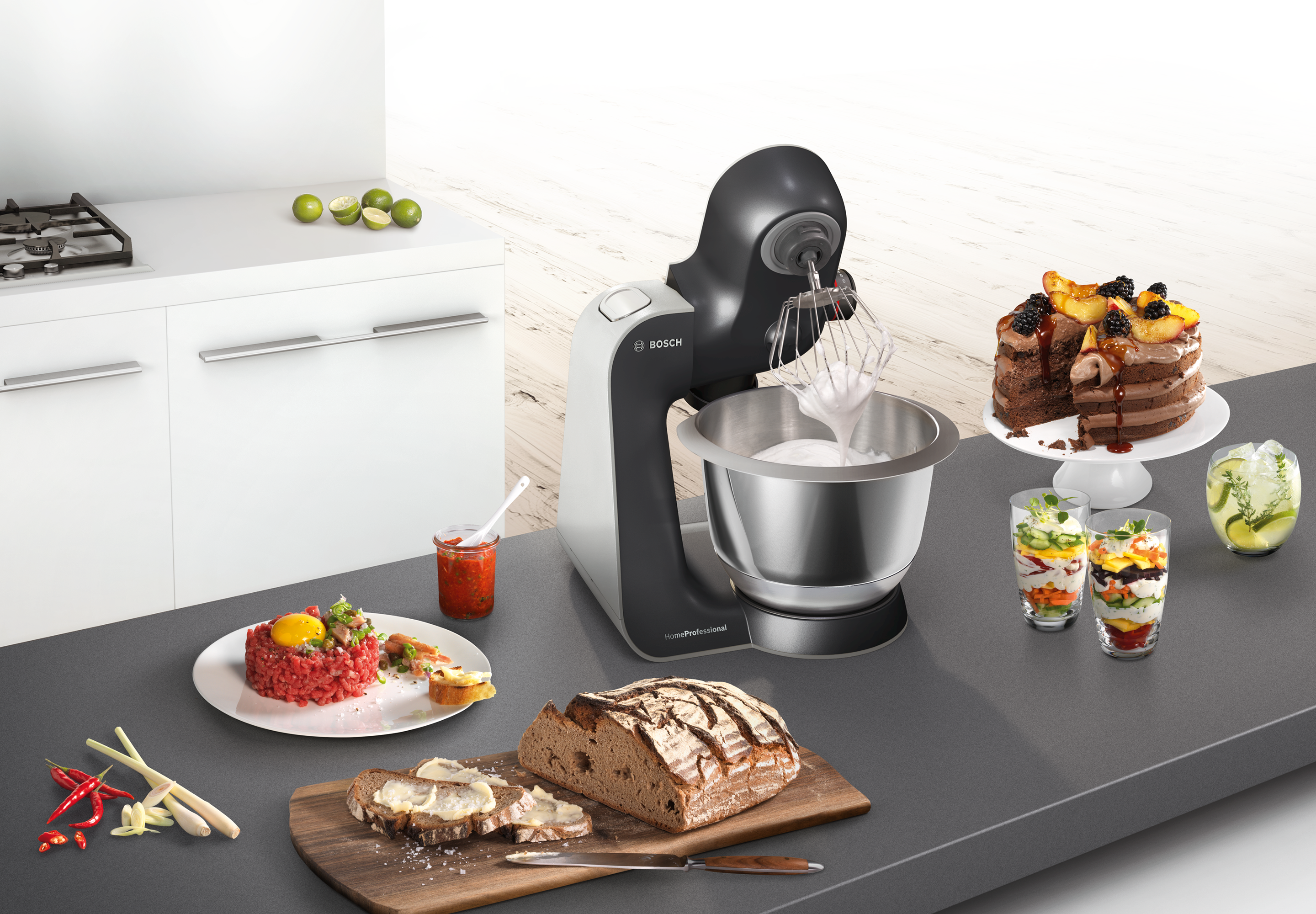 A kitchen scene with a Bosch mixer, cake, food samples and bread on a countertop. White cabinets and stove.