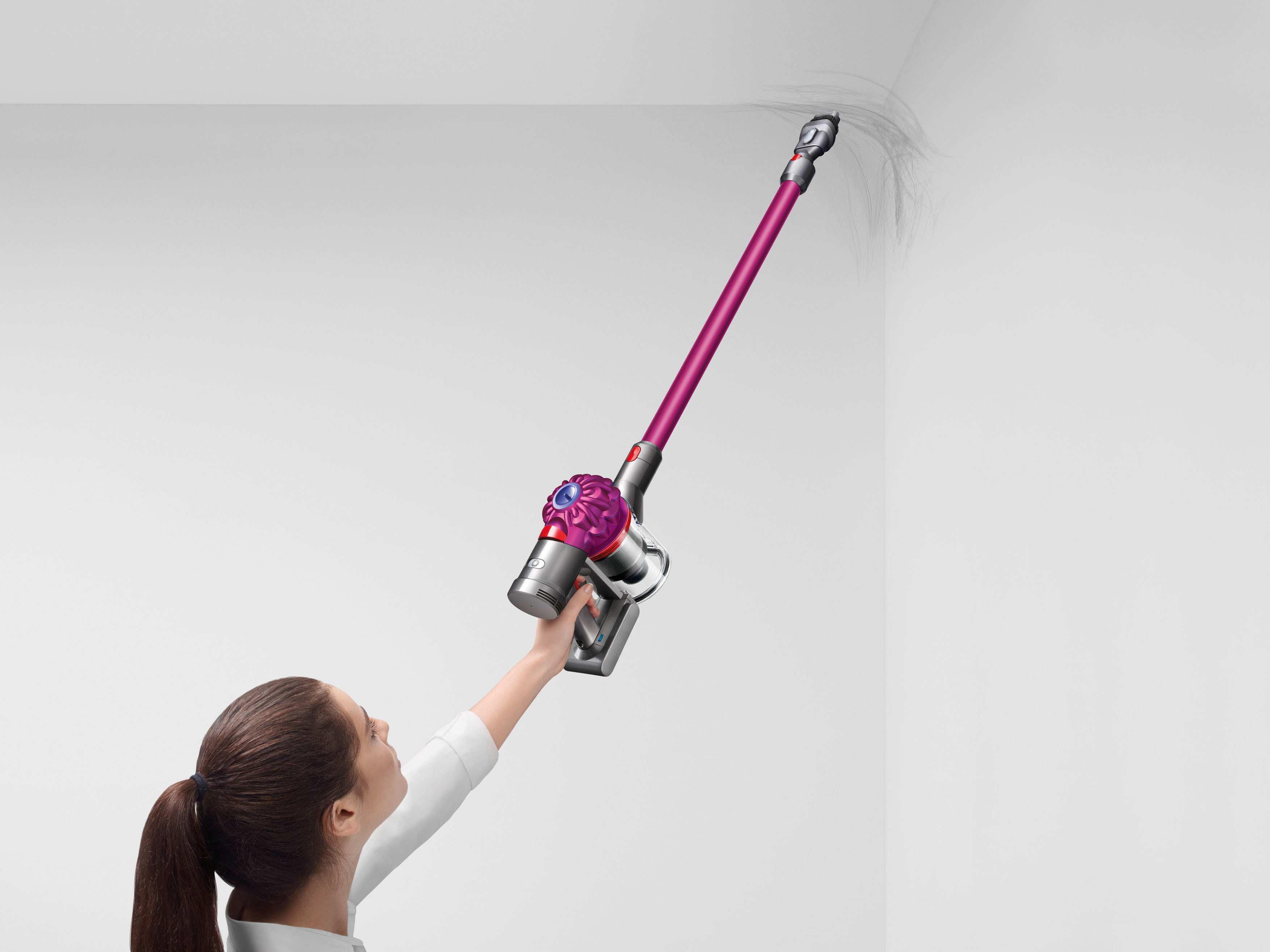 Eine Frau benutzt einen pinkfarbenen und silbernen Staubsauger an einer weißen Wand. Staub wird aufgesaugt.