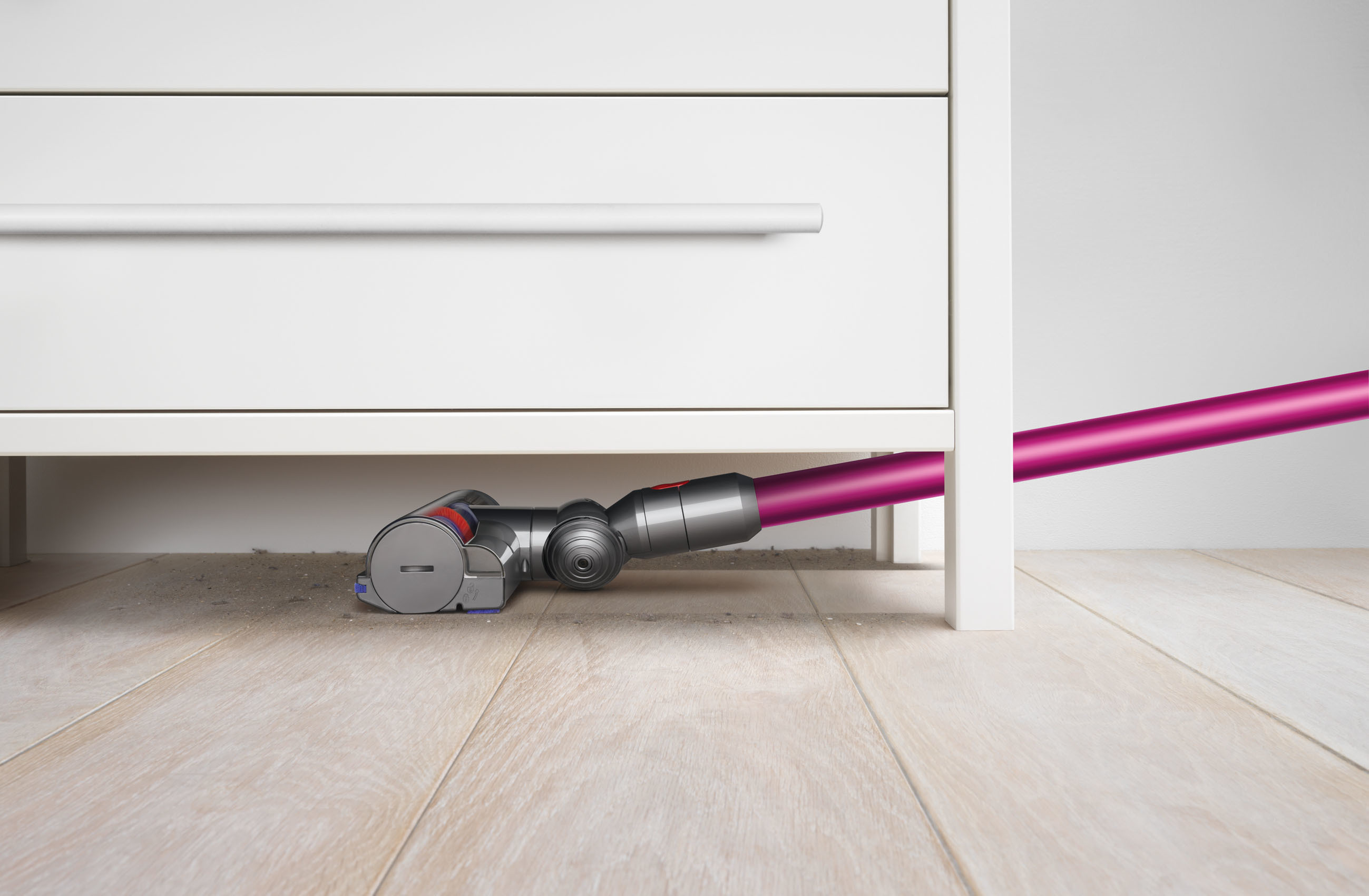 Ein Staubsauger mit lila Rohr reinigt unter einem weißen Schrank auf einem Holzboden.
