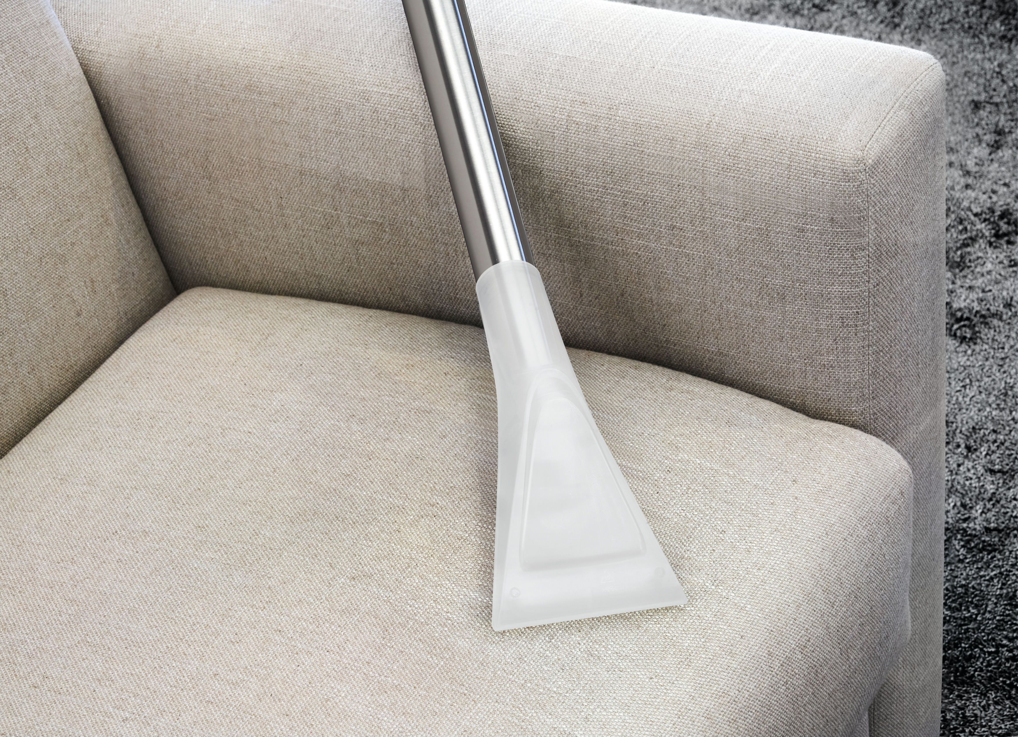 Ein Staubsaugerkopf reinigt ein Sofa. Der Kopf ist transparent, das Sofa ist beige.