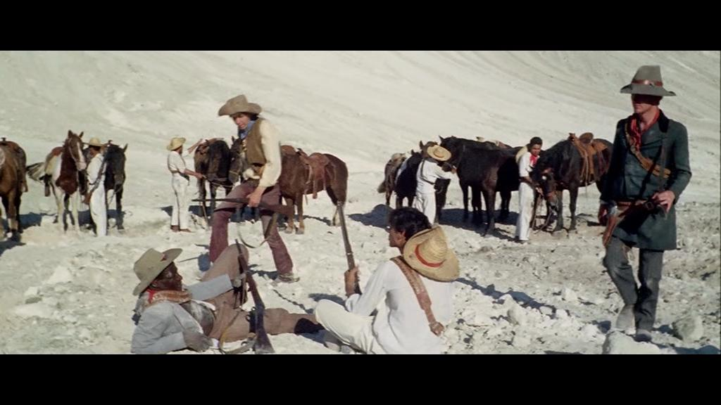 Cowboys in einer Wüstenszene, einige sitzen, andere stehen in der Nähe von Pferden.