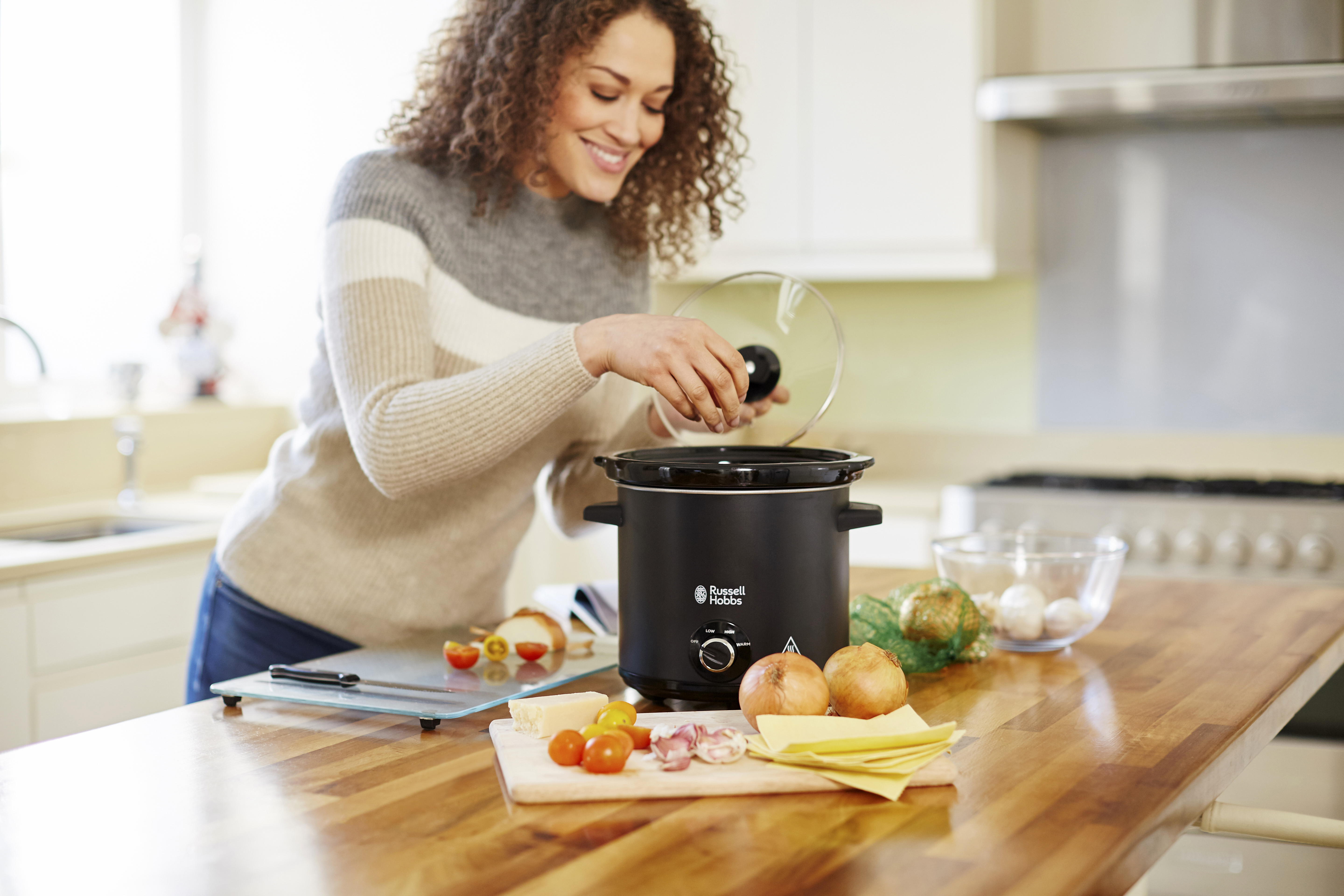 Vrouw voegt ingrediënten toe aan een zwarte slowcooker op een houten aanrecht. Groenten en kaas in de buurt.