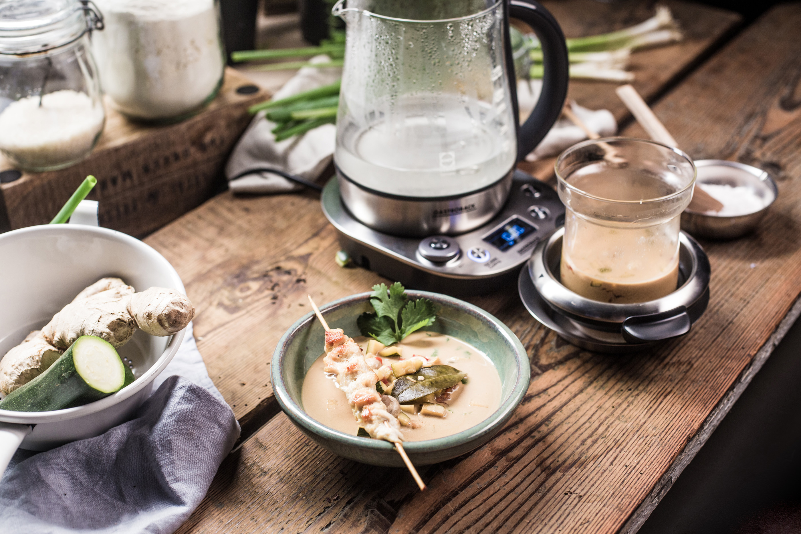 Tisch mit Suppe, Ingwer, einem Wasserkocher und Zutaten. Ein Spieß mit Essen liegt in der Suppe.